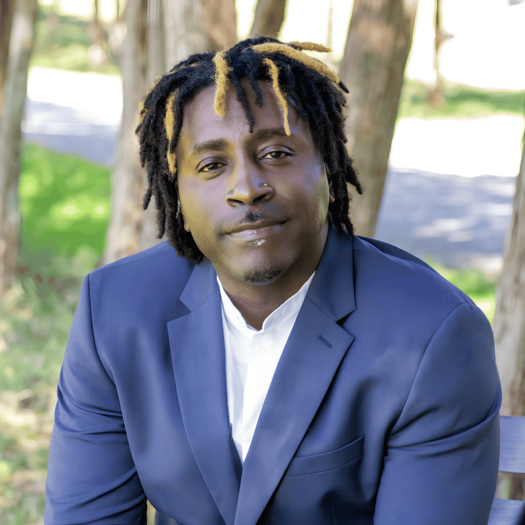 A man with dreadlocks with blonde tips wears a navy blazer over a white shirt, seated outdoors among trees | ReEvolve Therapy