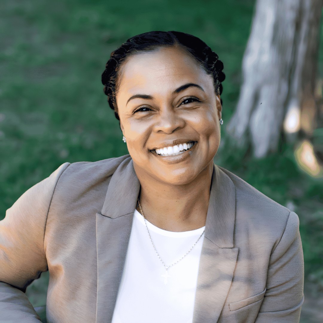 A smiling woman with braided hair wears a tan blazer and a cross necklace, standing outdoors in front of greenery | ReEvolve Therapy
