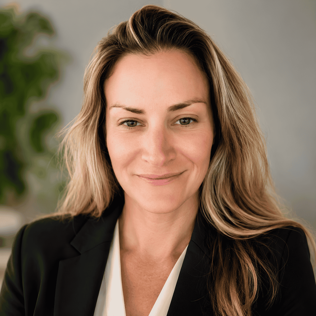 A smiling woman with long blonde hair wears a black blazer over a white top, photographed against a softly blurred indoor background with greenery | ReEvolve Therapy