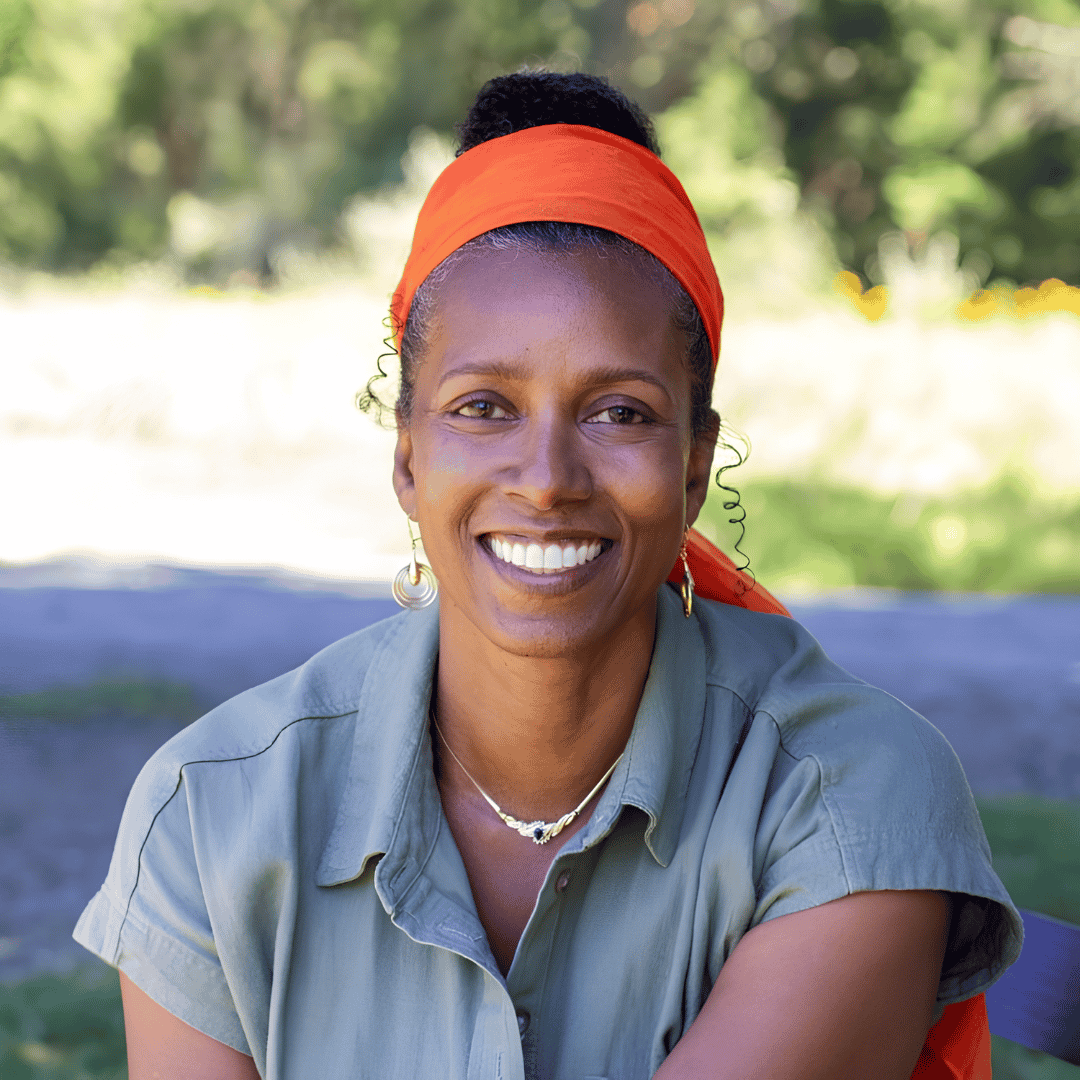 A smiling woman wearing an orange headwrap and hoop earrings sits outdoors in a sage green shirt, with greenery in the background | ReEvolve Therapy
