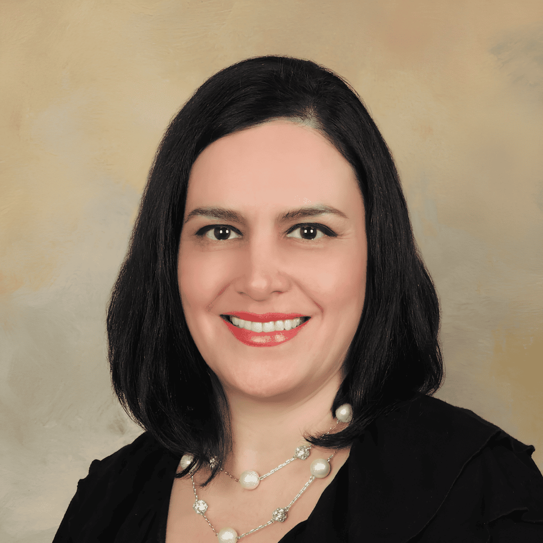 A smiling woman with dark shoulder-length hair wears a black top with a layered pearl necklace, photographed against a warm textured background | ReEvolve Therapy