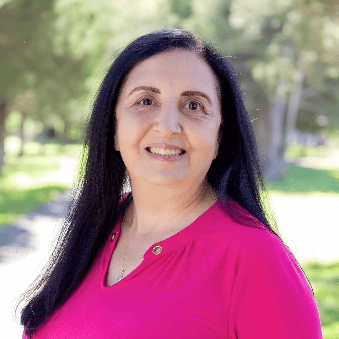 A smiling woman with long dark hair wears a bright pink top, photographed outdoors with trees and a path in the background | ReEvolve Therapy