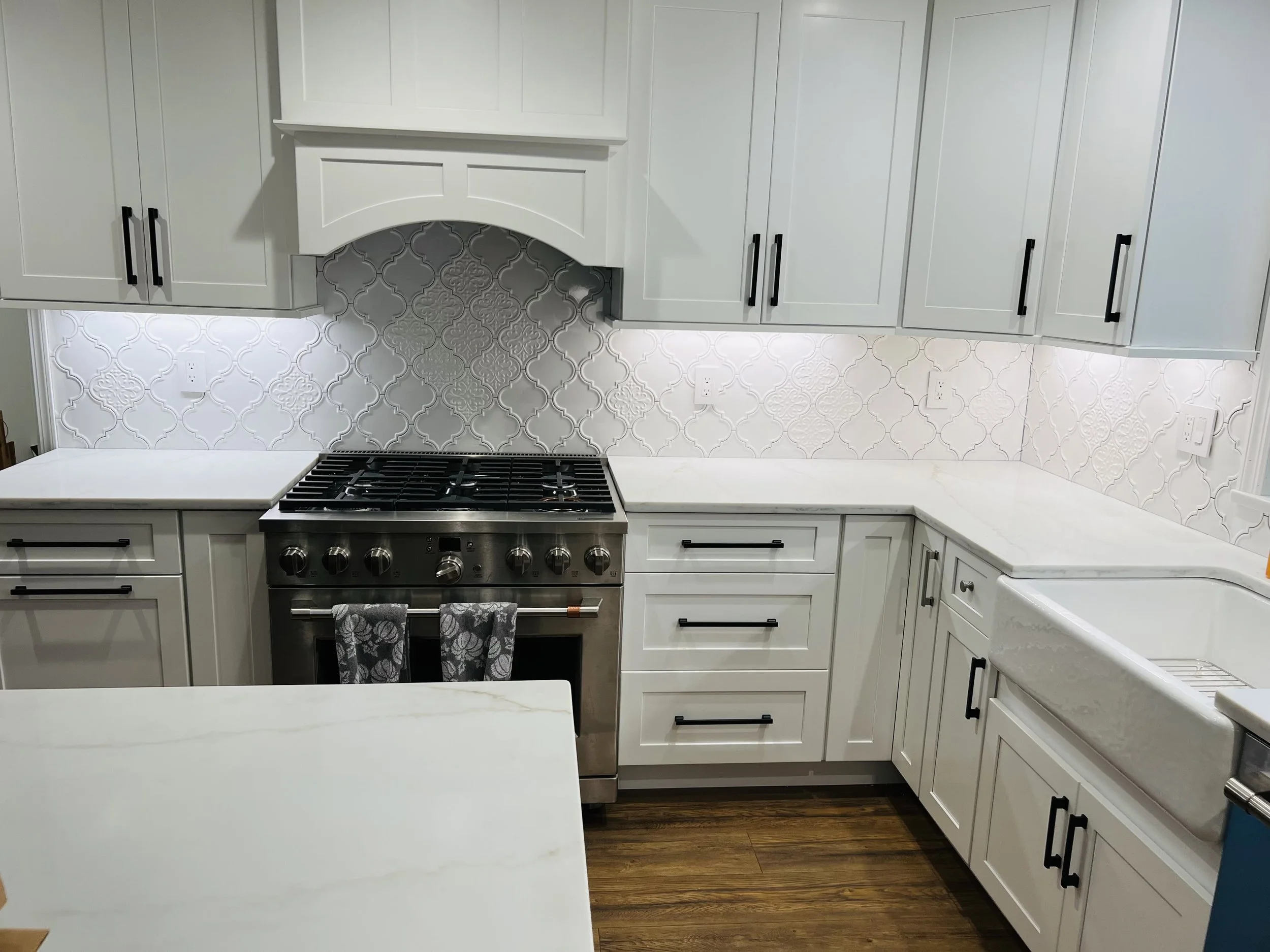 A modern white kitchen with a backsplash featuring decorative tiles, white cabinets with black handles, a stainless steel stove, and a white countertop.
