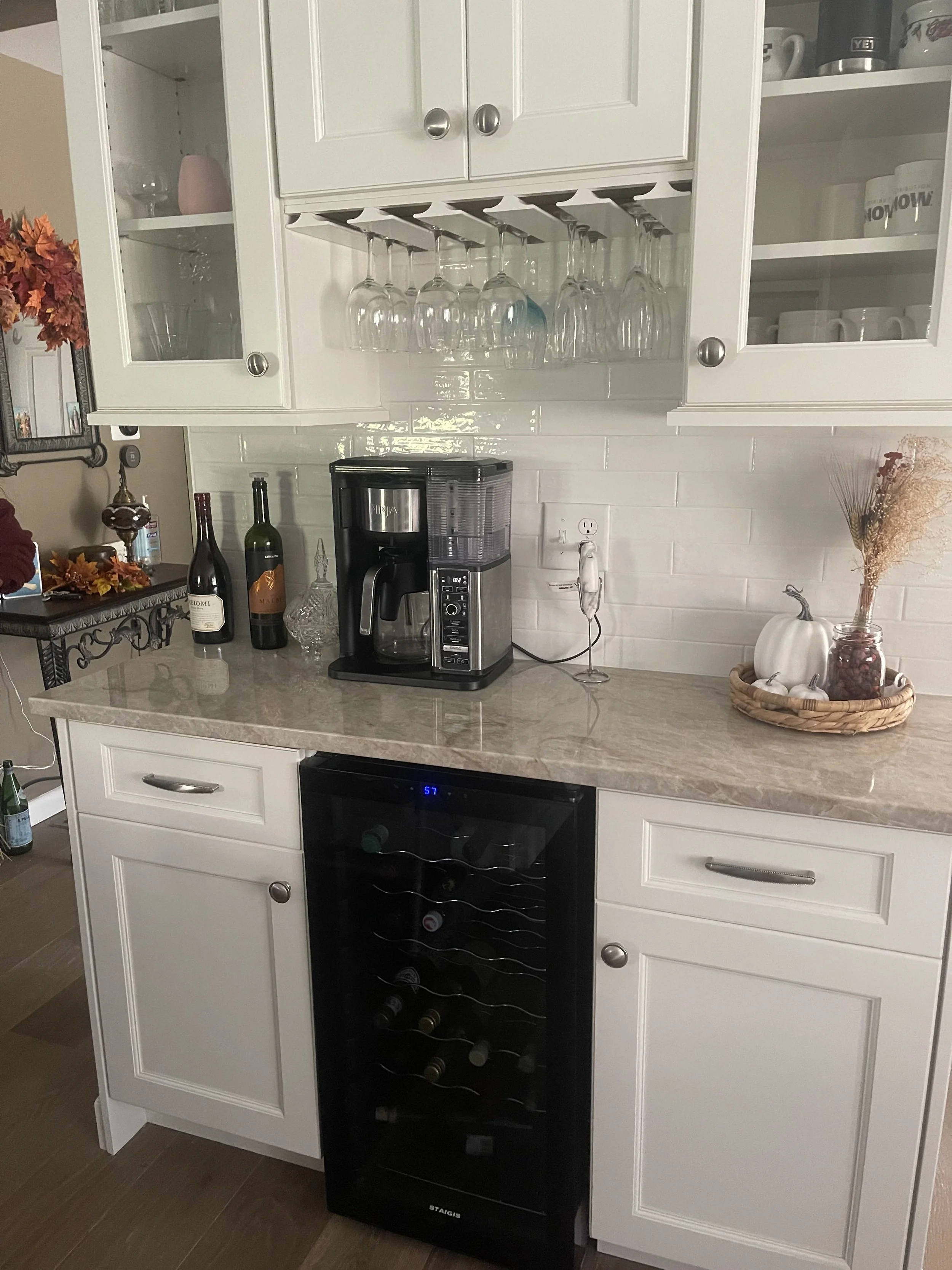 White kitchen cabinet with glass doors and wine glass rack, coffee maker on beige countertop, decorative pumpkins and fall foliage, bottles of wine, and a wine cooler underneath.