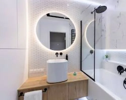 Modern bathroom with a round illuminated mirror, white sink on a wooden vanity, black fixtures, and a glass shower door.
