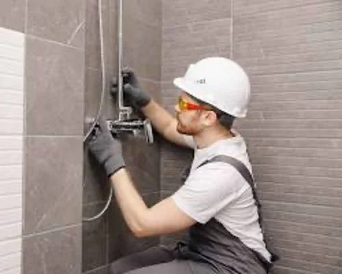 A construction worker wearing a white safety helmet, safety glasses, and gloves, installing or repairing plumbing fixtures in a tiled bathroom or shower area.