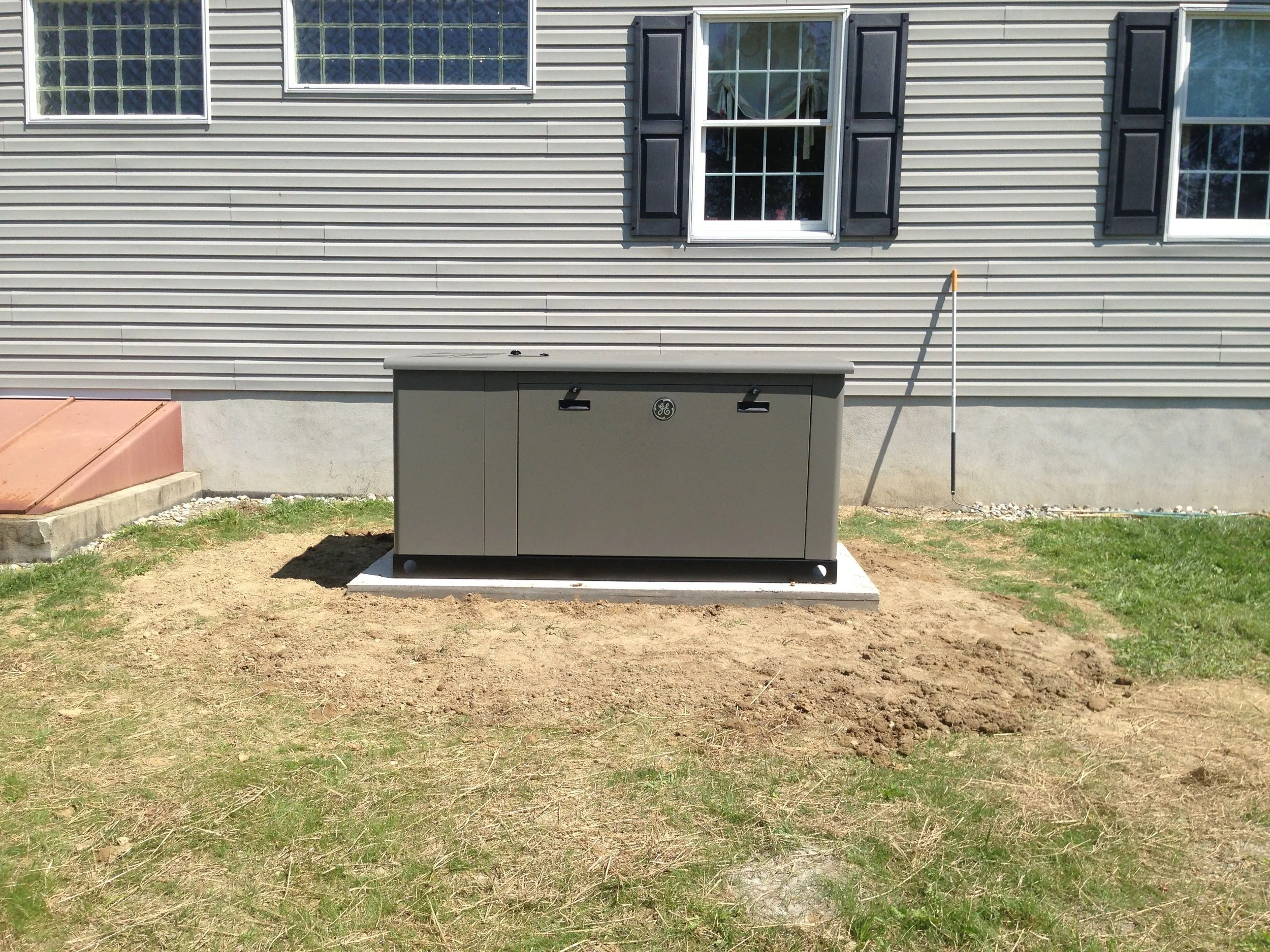 View of the side of a house with beige vinyl siding, three windows with black shutters, a gray electrical box, and a white pole on the ground. The yard has partly grass and bare dirt.