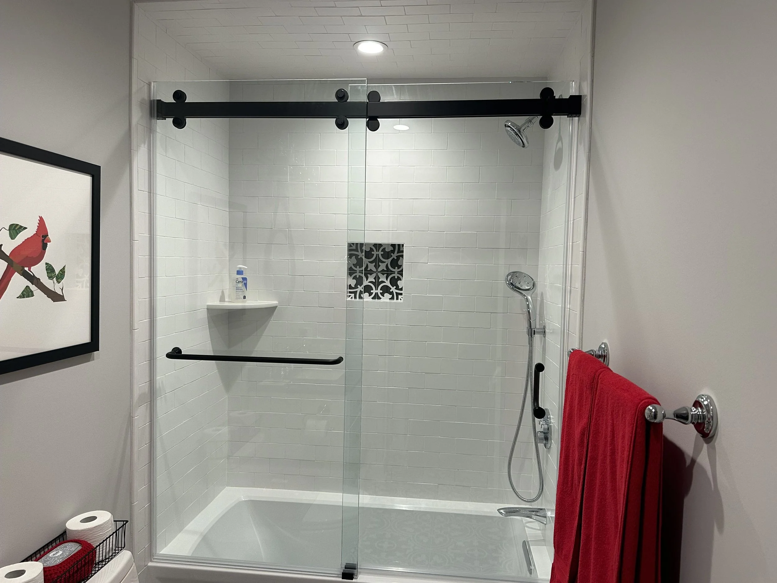 A modern bathroom shower with a glass door, white subway tile walls, and a black sliding barn door hardware. Inside the shower, there is a handheld showerhead and a small shelf with soap and toiletries. A framed picture of a red cardinal bird is partially visible on the wall outside the shower. Red towels are hanging on a silver towel bar.