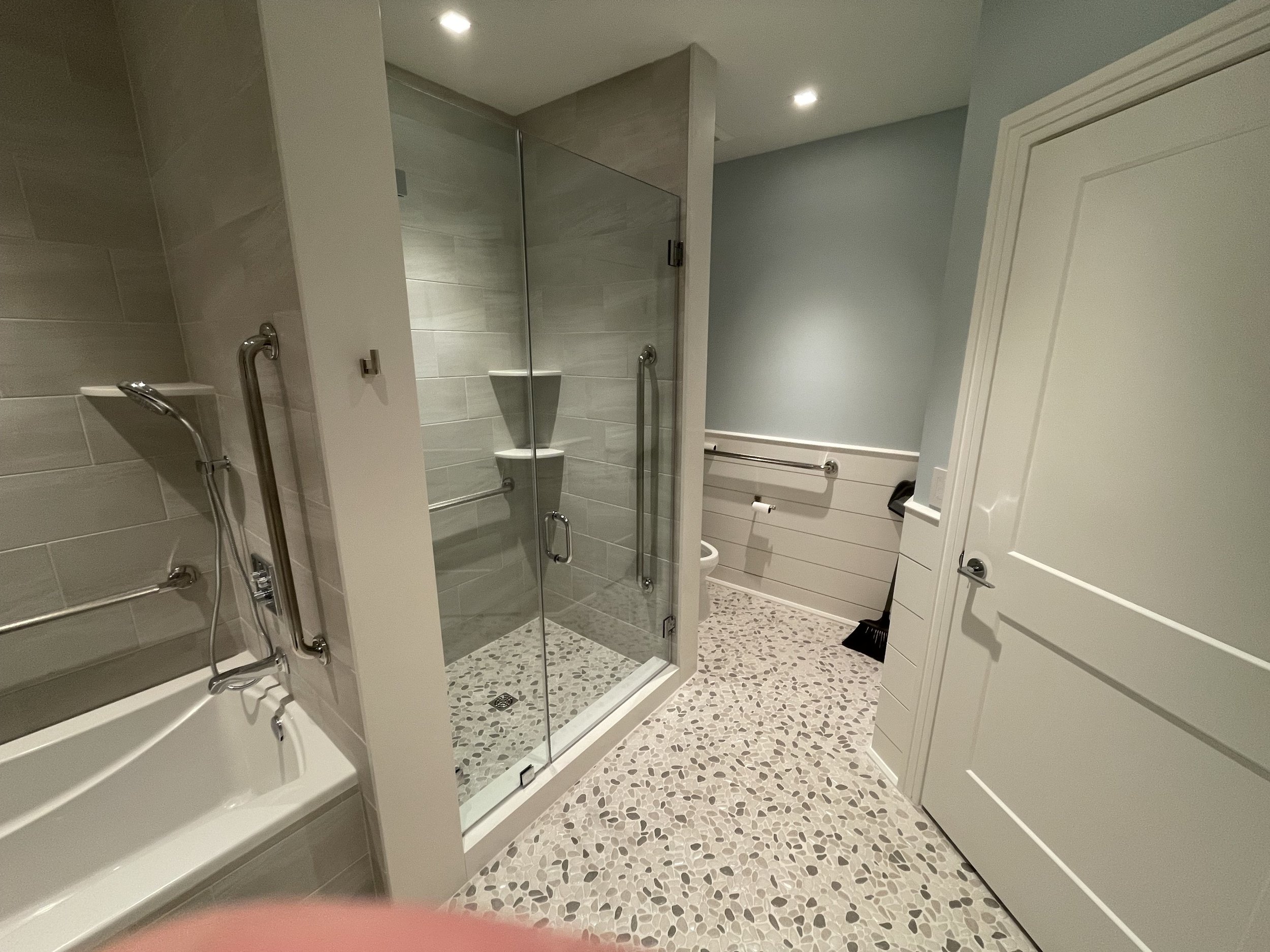 A modern bathroom with a shower enclosure featuring glass doors, gray wall tiles, a handheld shower head, and a built-in corner shelf. The bathroom has a terrazzo tiled floor, a white door, a white cabinet, and a bidet.