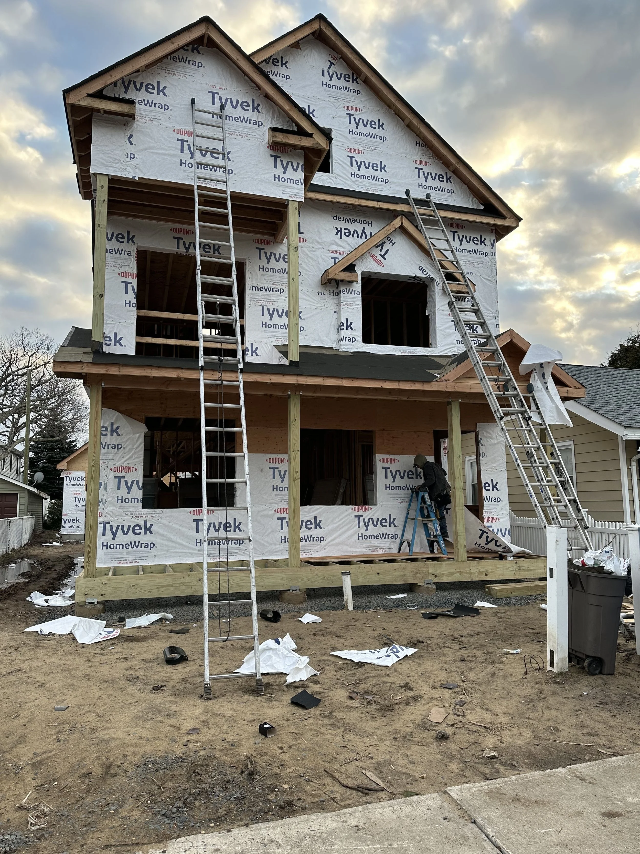 A two-story house under construction with wooden framing and white weather-resistant wrap, scaffolding, ladders, and construction workers present, on a dirt lot.