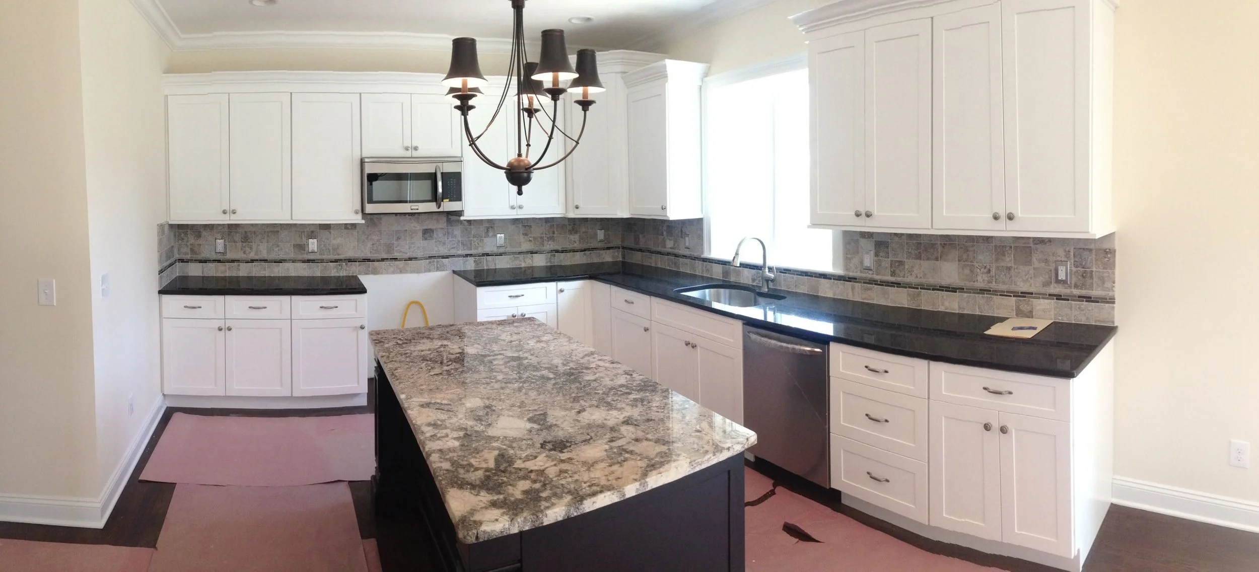 A kitchen with white cabinets, black granite countertops, a decorative tile backsplash, and a central kitchen island with a granite top. There is a chandelier hanging from the ceiling.