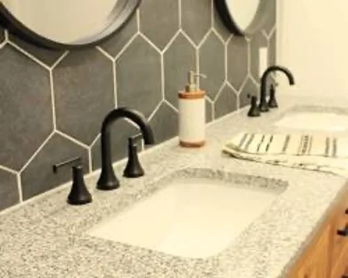 Bathroom countertop with two sinks, black faucets, soap dispenser, and striped towel, with hexagonal tile backsplash.