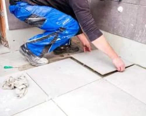 Person installing tiles on a floor, kneeling and using tools to place tiles.