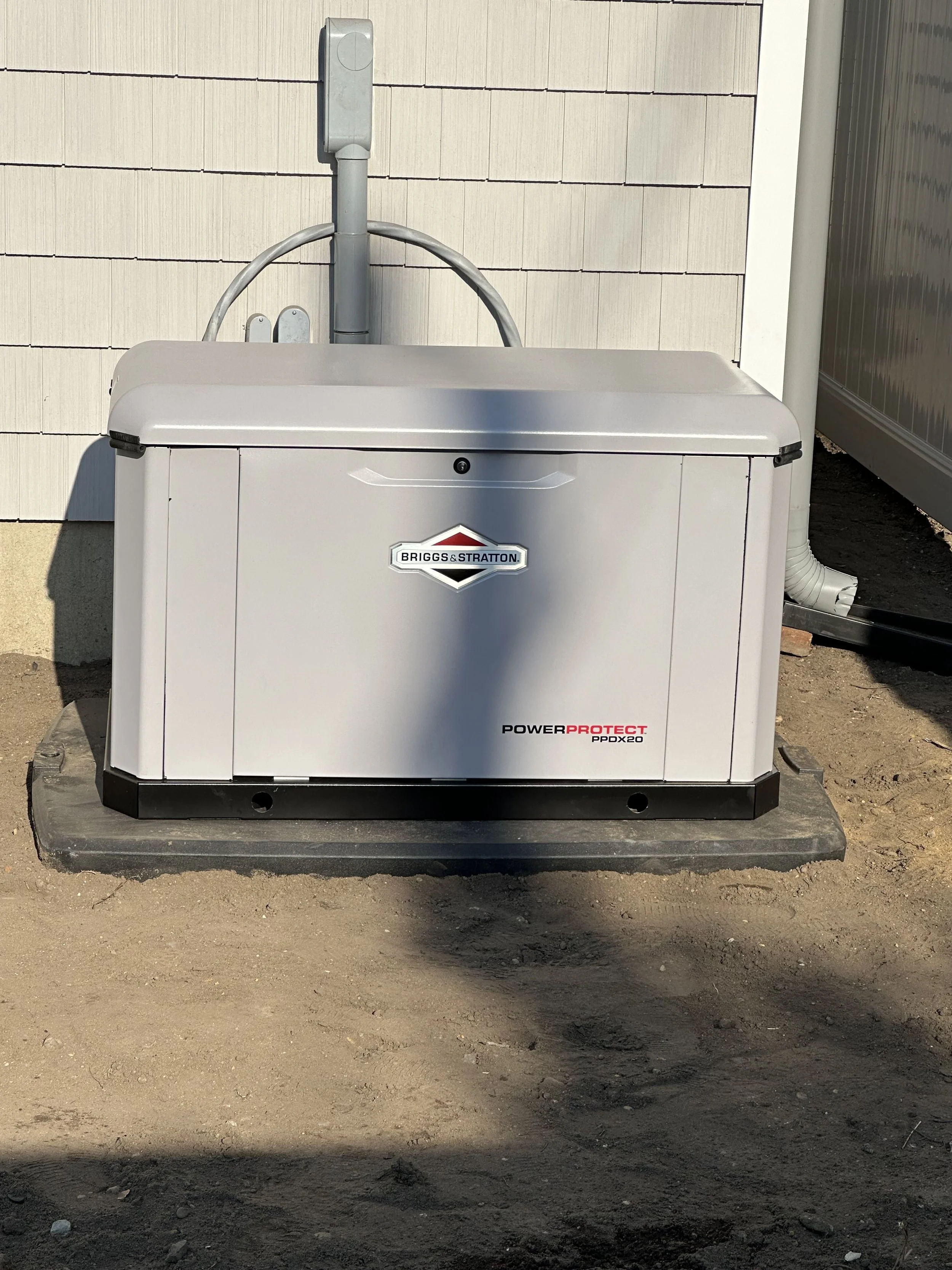 A gray Briggs & Stratton PowerProtect generator on a concrete pad outside, with a background of a beige vertical siding wall and a section of a building on the right.