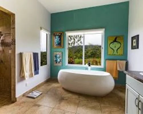 Bathroom with a white oval bathtub facing a large window, colorful paintings on the teal accent wall, with towels hanging on a wooden rack and a towel on a white cabinet.
