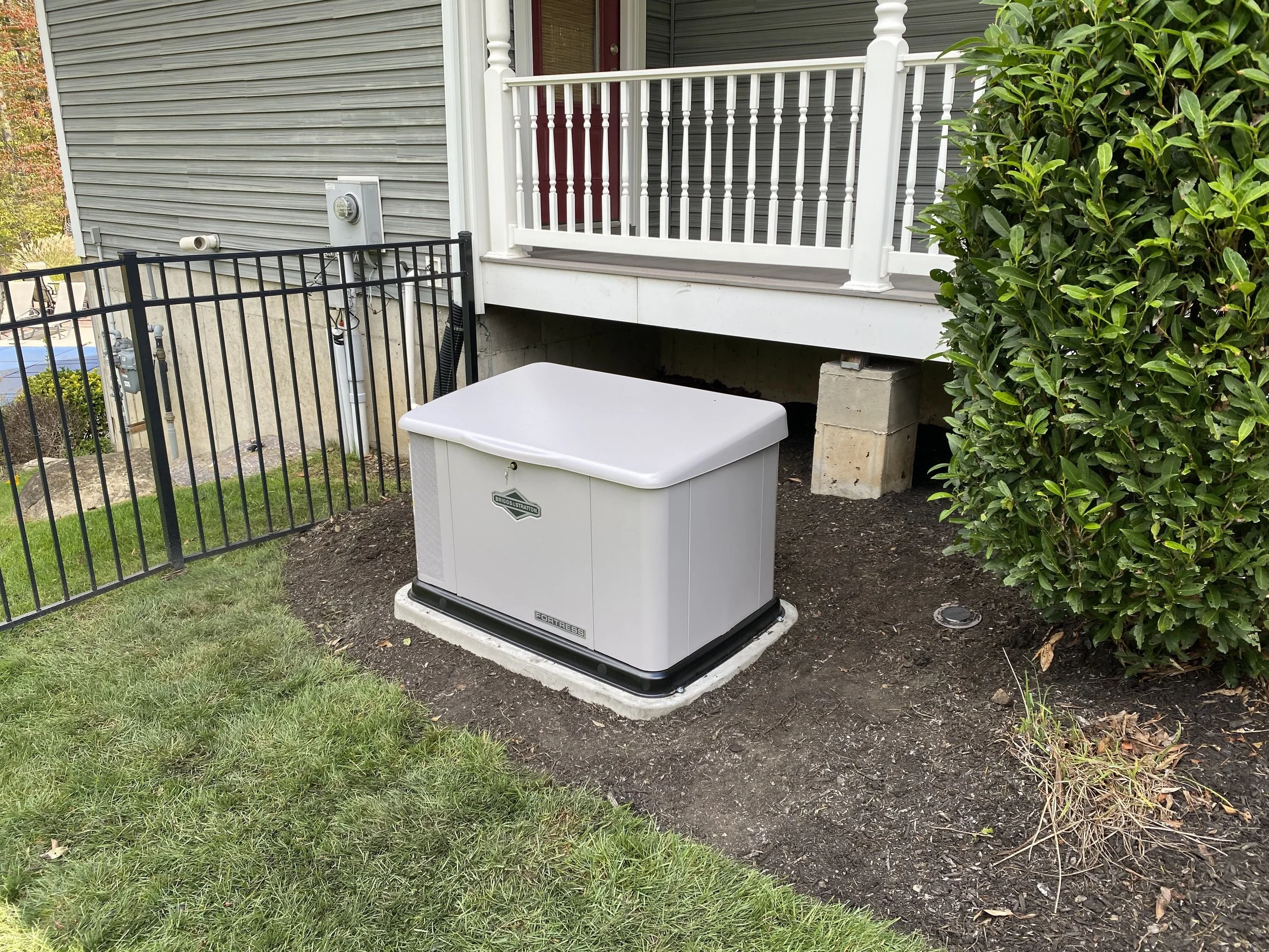Backyard view showing a small porch with white railing, a hedge on the right, a black metal fence on the left, a gray house exterior with siding, a utility box, and a white outdoor air conditioning unit on a concrete pad.