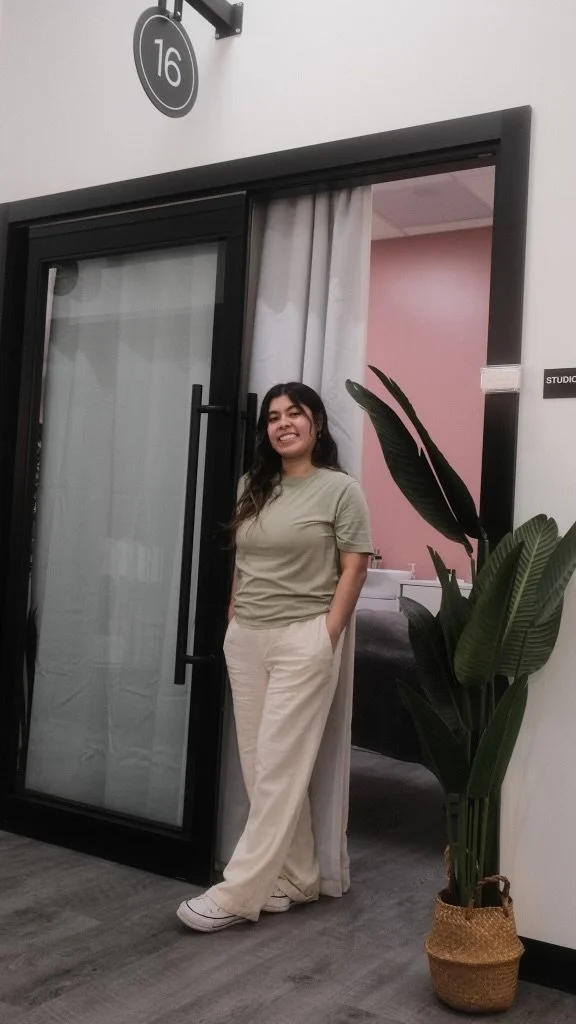 Person smiling and standing at the entrance of a studio or office with sign number 16, next to a plant, wearing casual clothes.