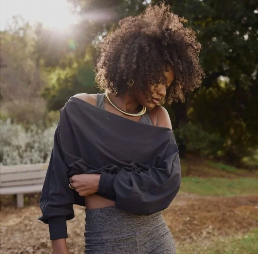 A woman with curly hair wearing a black jacket off her shoulders, a gray top, and matching gray pants, standing outdoors in sunlight with trees and a park bench in the background.