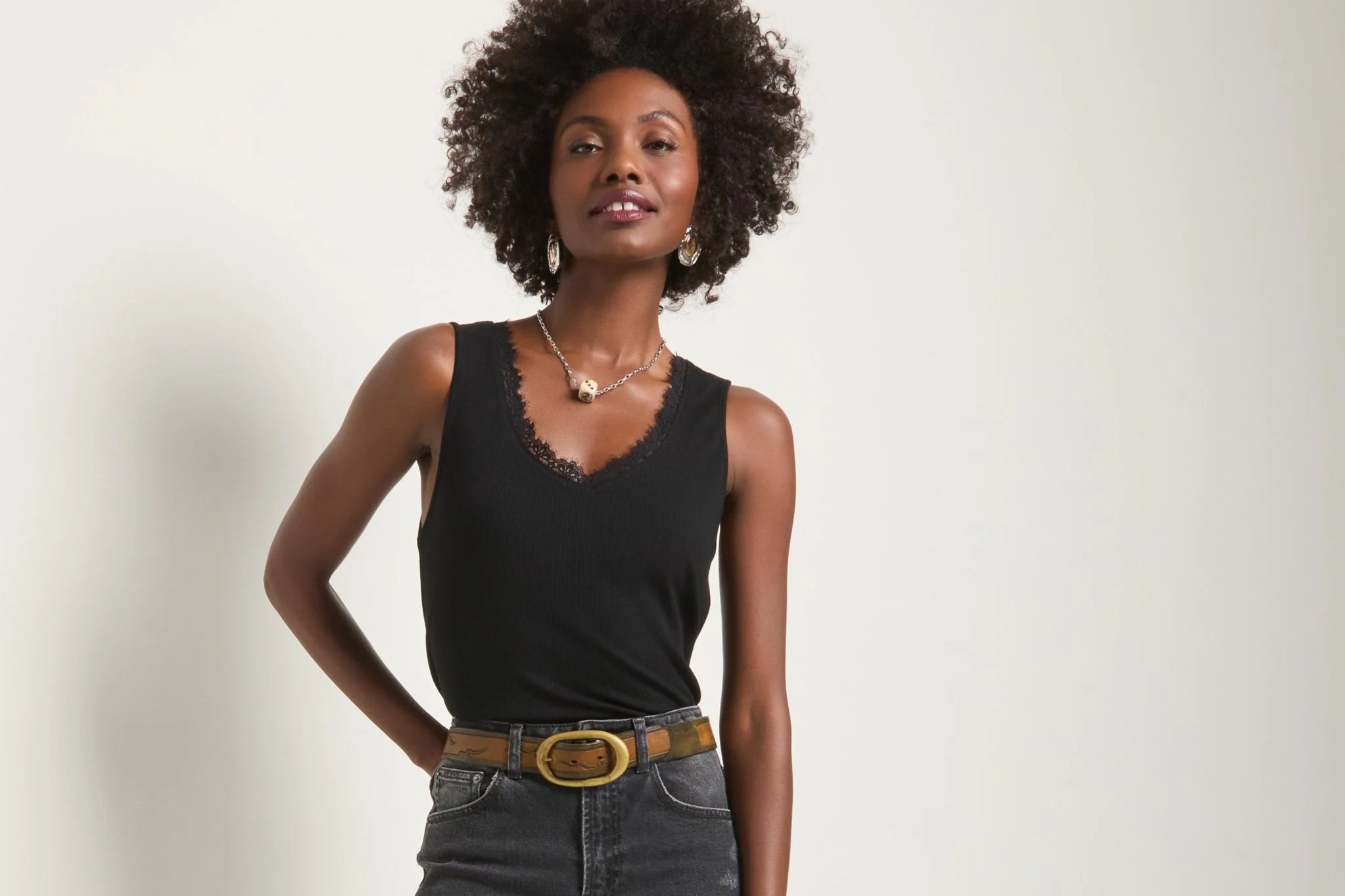 A young woman with curly black hair wearing a black tank top with lace detail, silver jewelry including earrings and a chain necklace with a skull pendant, and high-waisted jeans with a brown belt, standing against a plain white background.