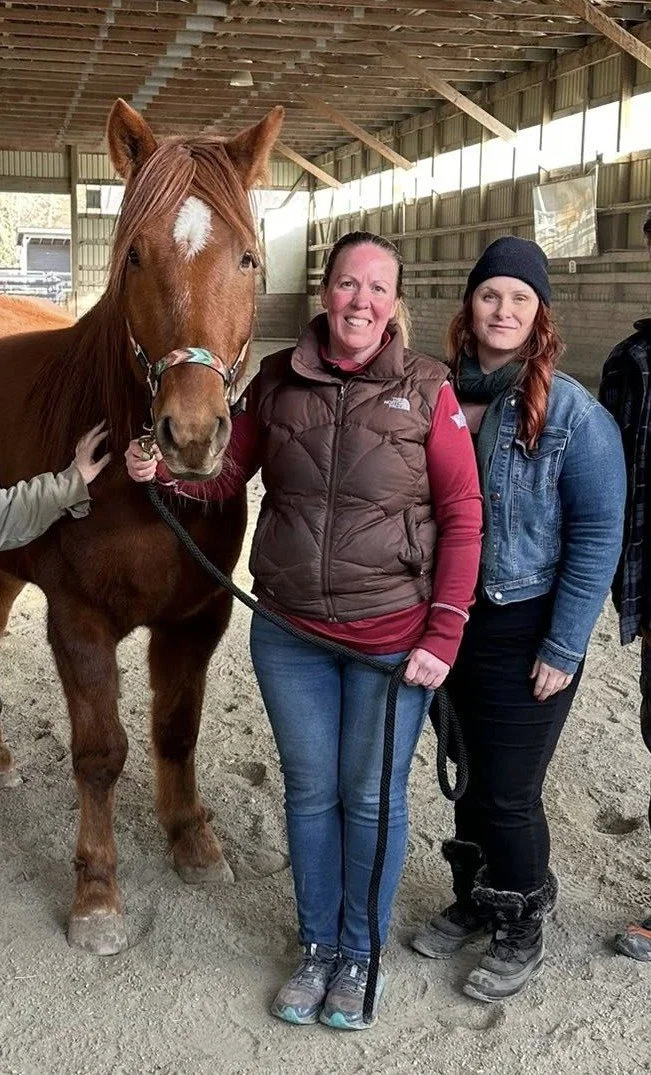 Two women sitting close together on outdoor white bench, hugging and smiling, with green grass, plants, and trees in the background.