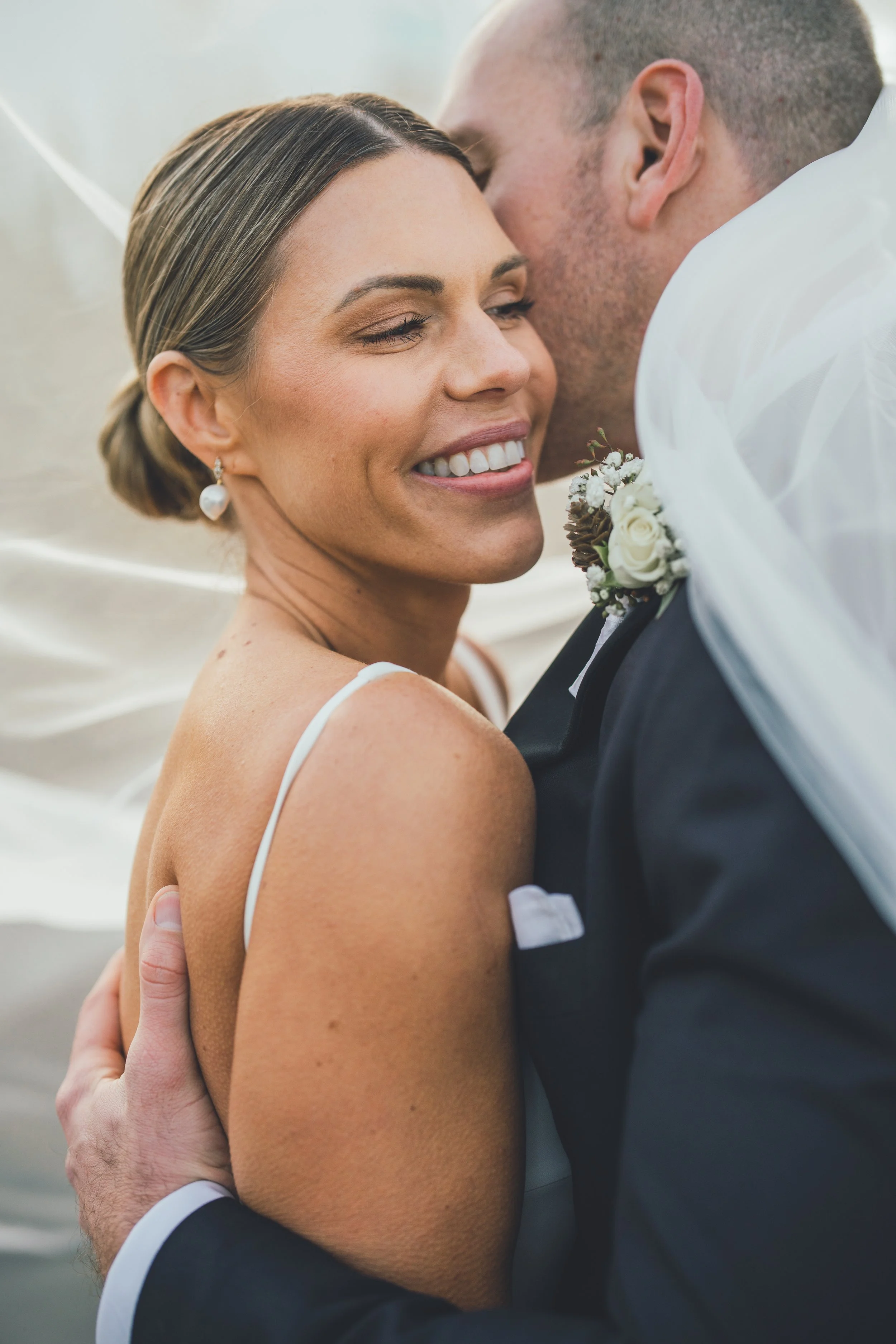A newlywed couple shares a kiss, smiling with eyes closed; the bride has a white dress and pearl earrings, and the groom is in a tuxedo with a boutonniere.