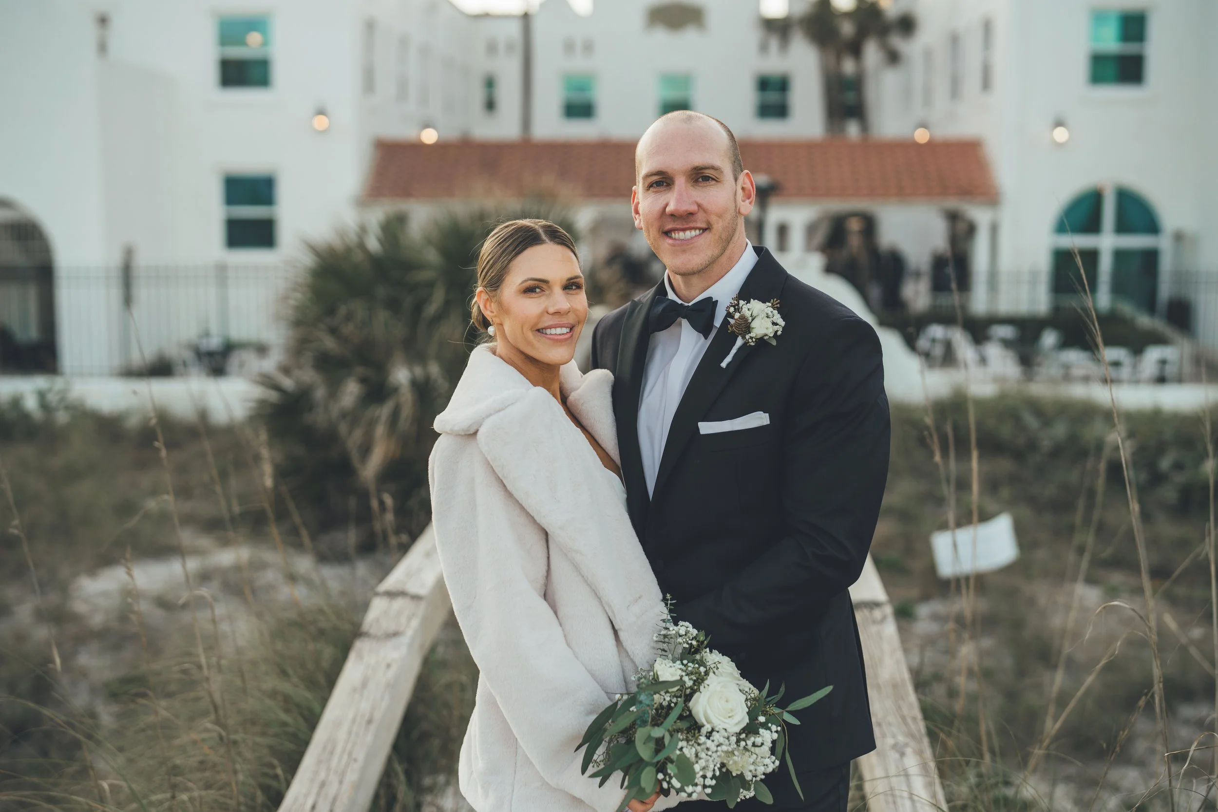 A newlywed couple smiling and standing close together outdoors on a wooden pathway, with the bride holding a bouquet of white roses and greenery, and the groom dressed in a black tuxedo with a bowtie, against a backdrop of beachside buildings and pla