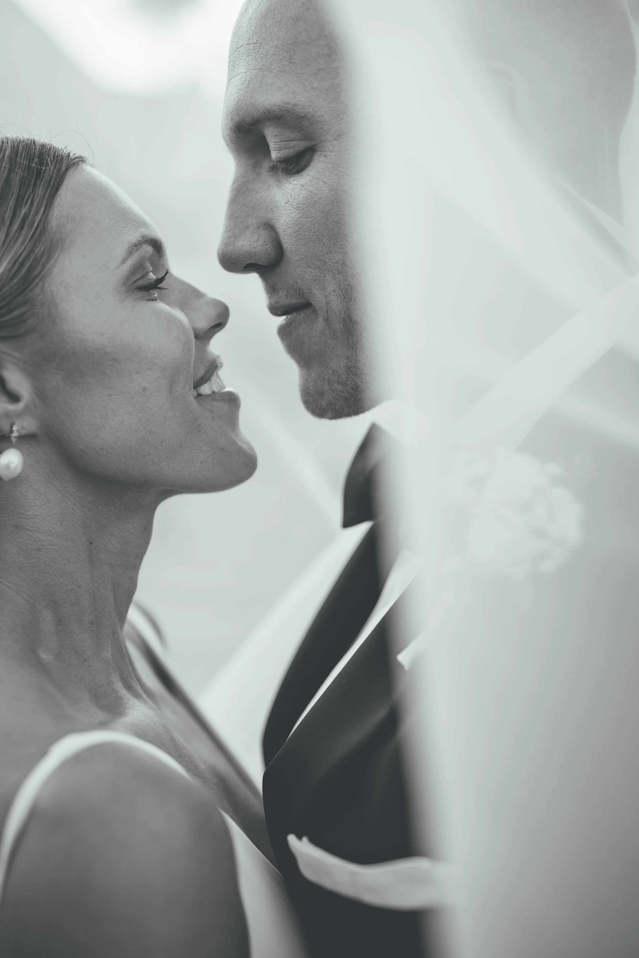 A close-up black and white photo of a bride and groom facing each other with their noses almost touching, smiling softly.