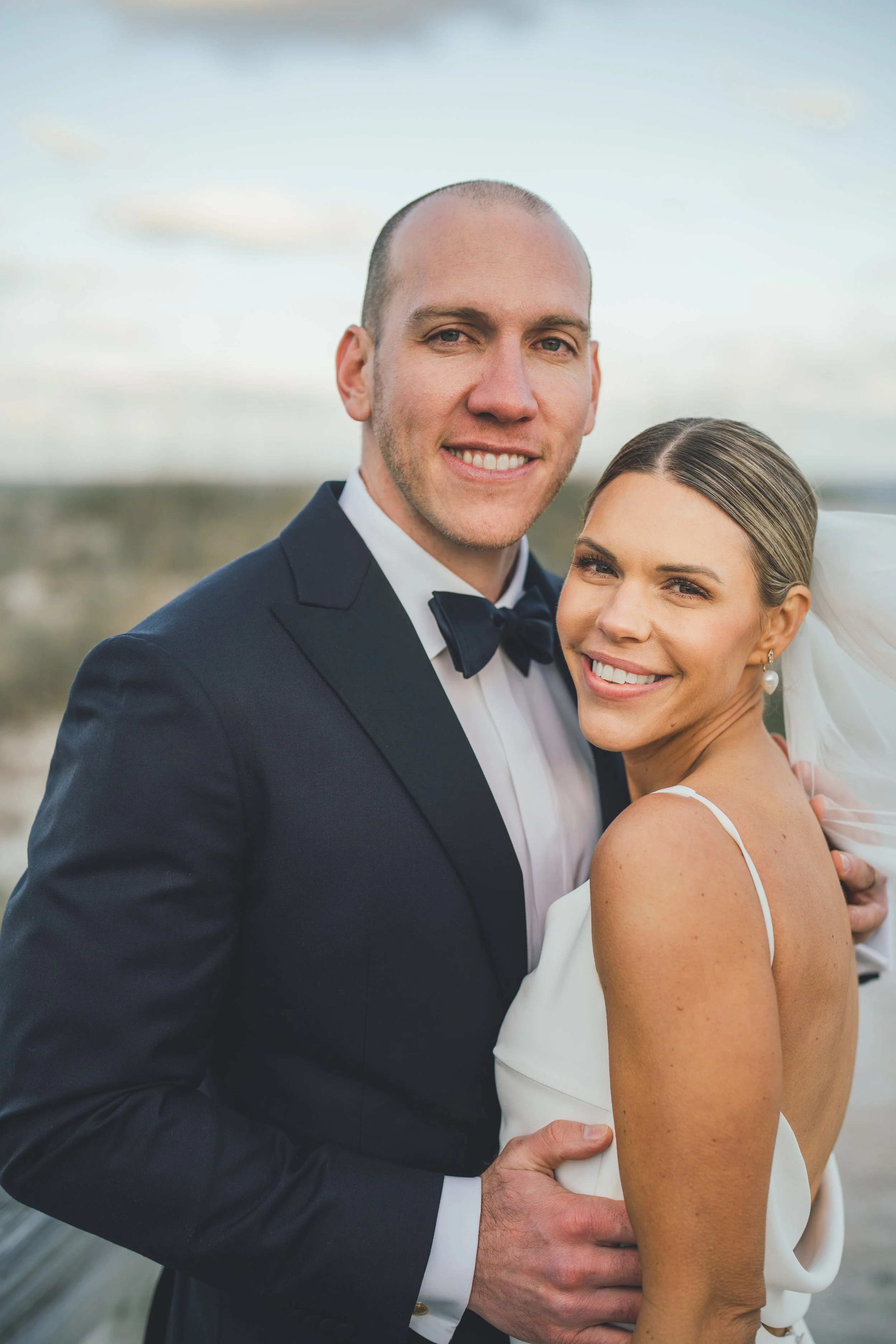 A happy newlywed couple smiling outdoors, with the groom in a black tuxedo and bow tie, and the bride in a white wedding dress with spaghetti straps, smiling and holding each other.