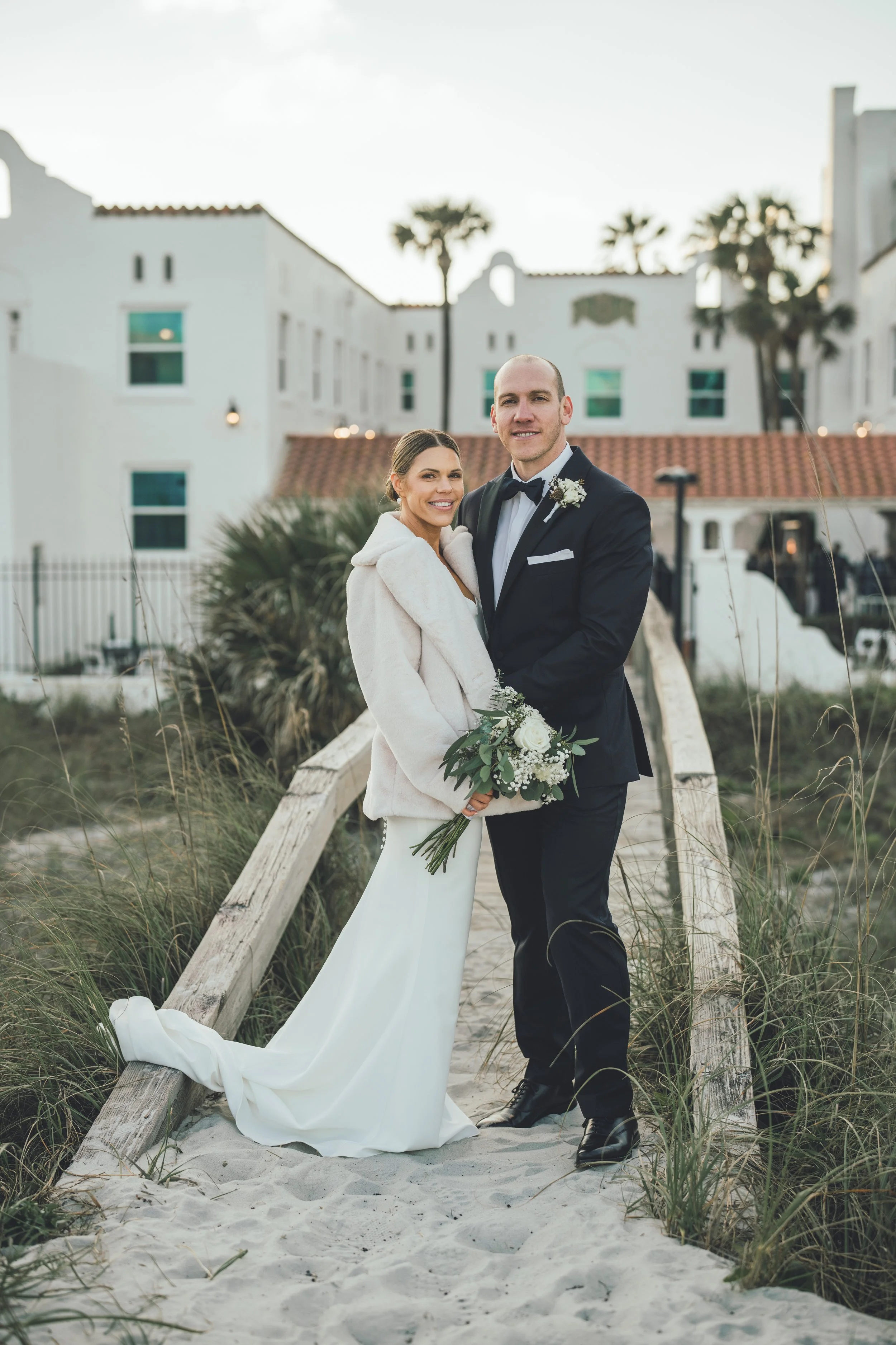 A bride and groom standing on a sandy path at the beach, smiling at the camera. The bride is holding a bouquet, wearing a white dress and a light-colored coat. The groom is dressed in a black tuxedo with a bow tie. There are buildings and palm trees 