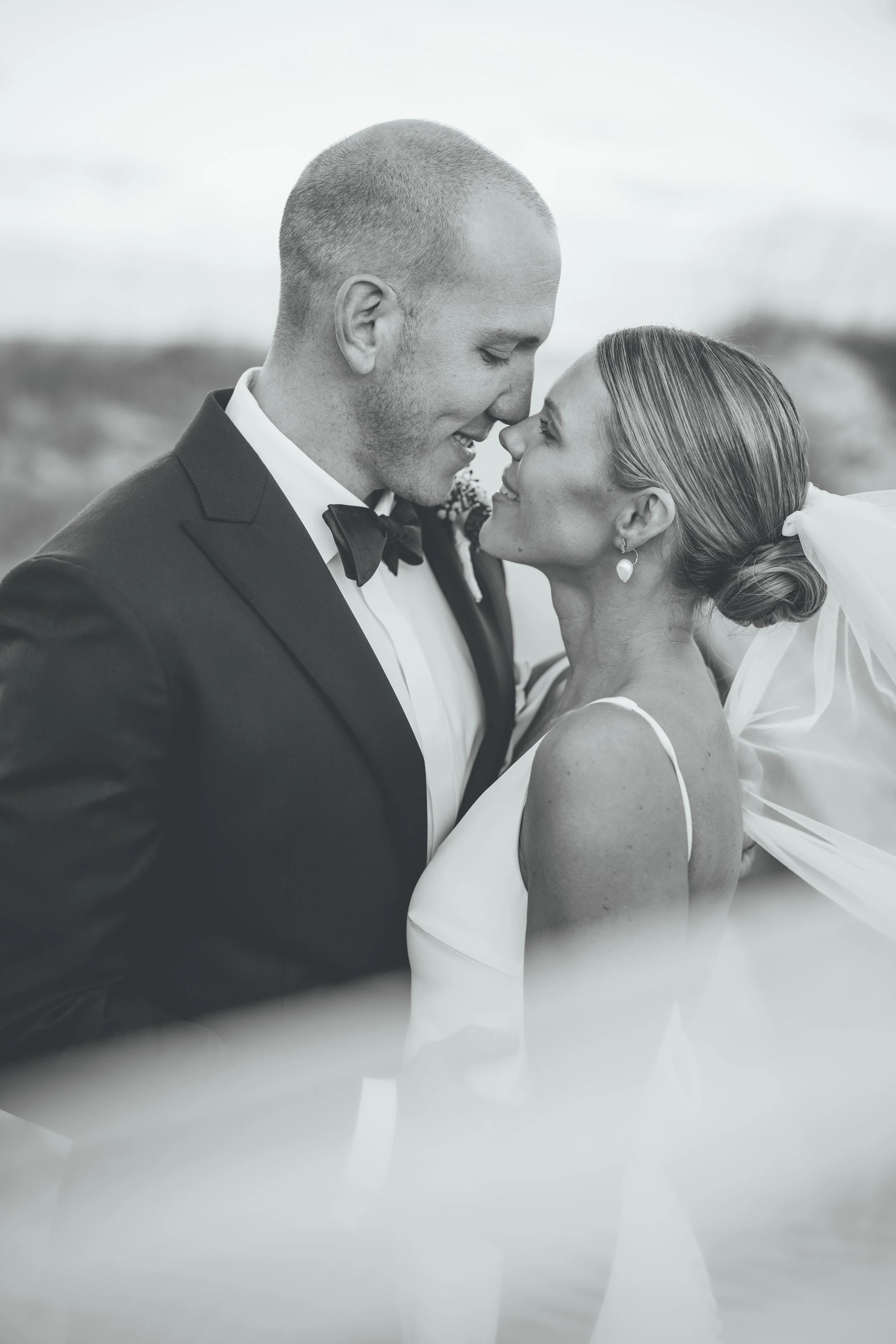 Black and white photograph of a newlywed couple embracing with their foreheads touching, outdoors during sunset.