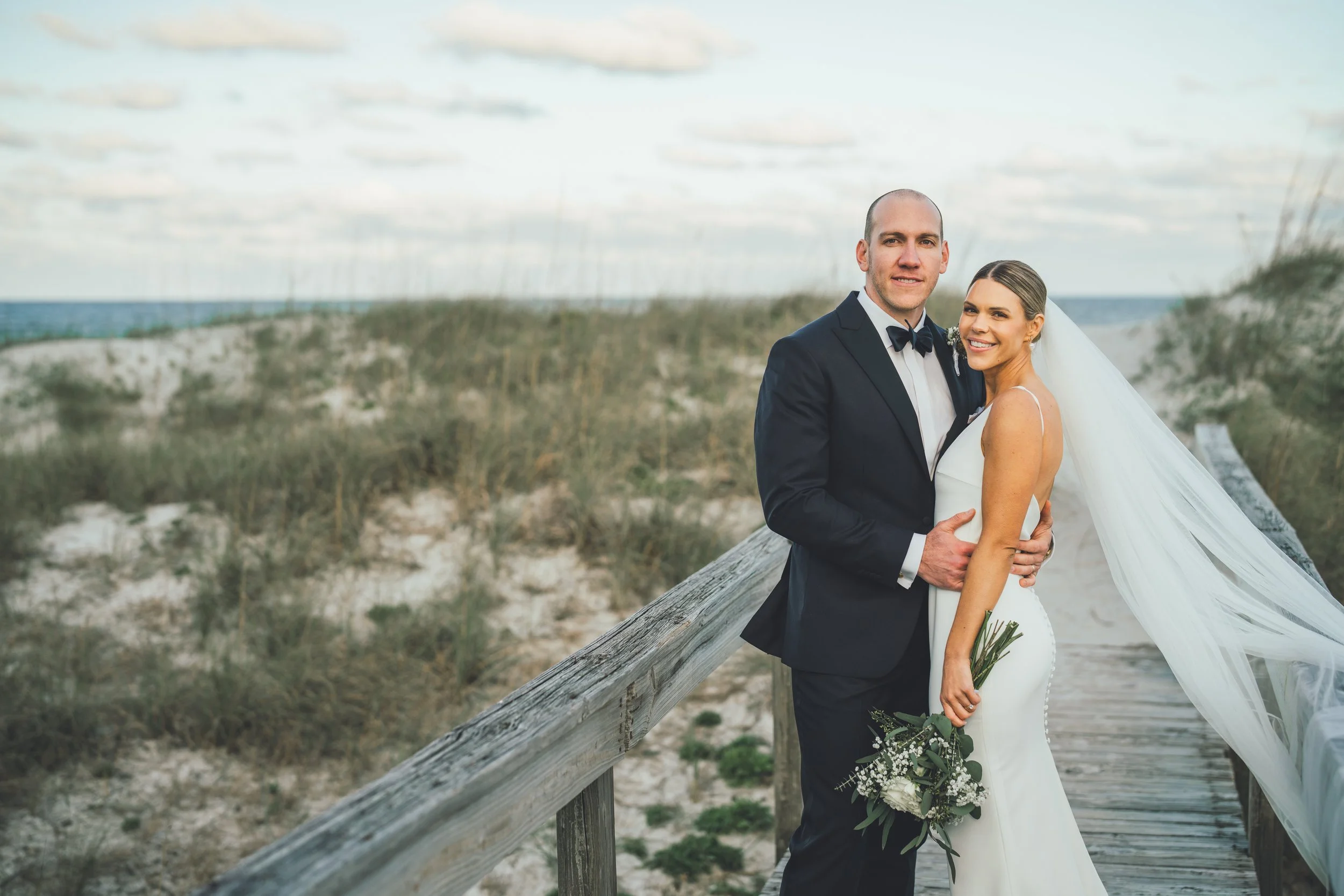 A happy bride and groom standing on a wooden boardwalk near the beach, holding each other and smiling, with the ocean and sky in the background.