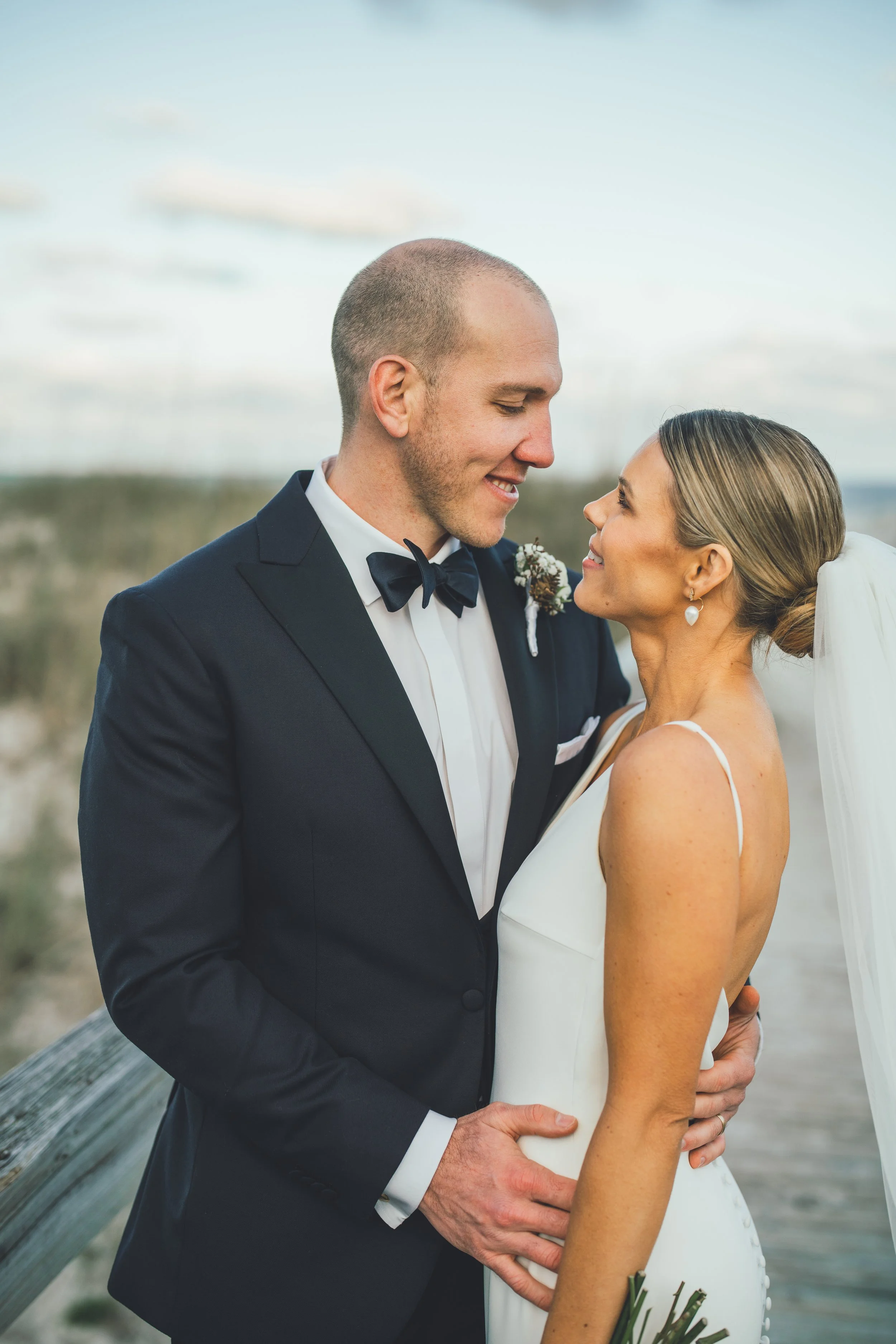 A newlywed couple stands close together outdoors on a wooden walkway, smiling and gazing into each other's eyes during sunset or late afternoon.