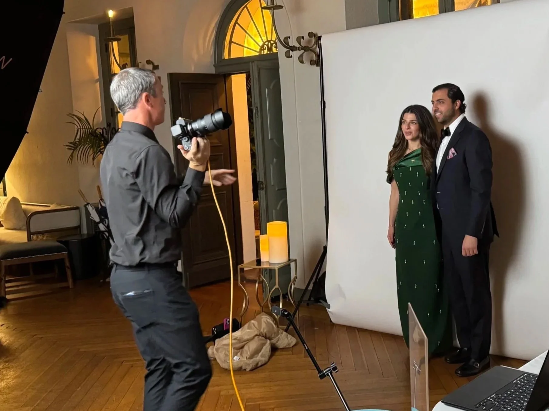 Photographer taking picture of a man and woman in formal attire standing in front of a white backdrop in a warmly lit room.