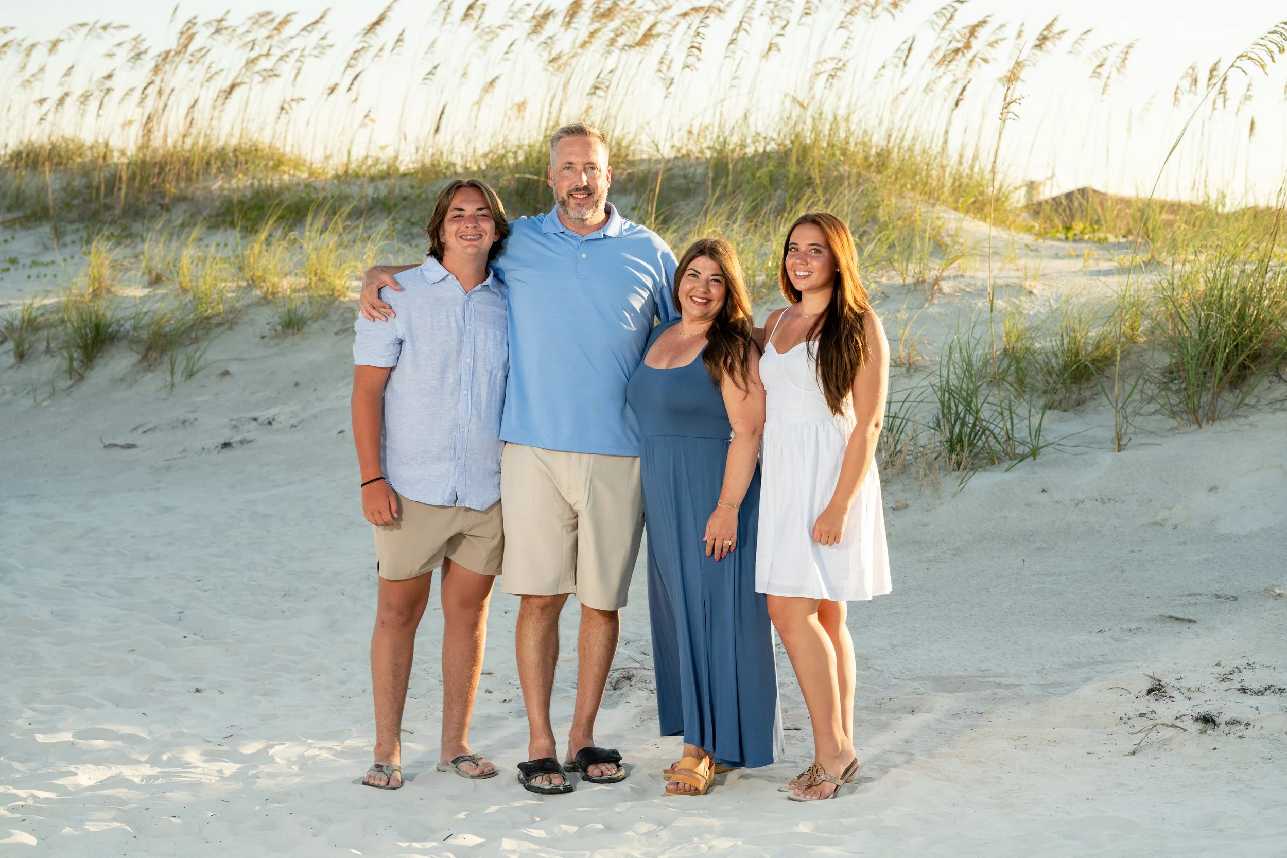 Family of four standing on sandy beach with grassy dunes and sunset in the background, smiling at the camera.