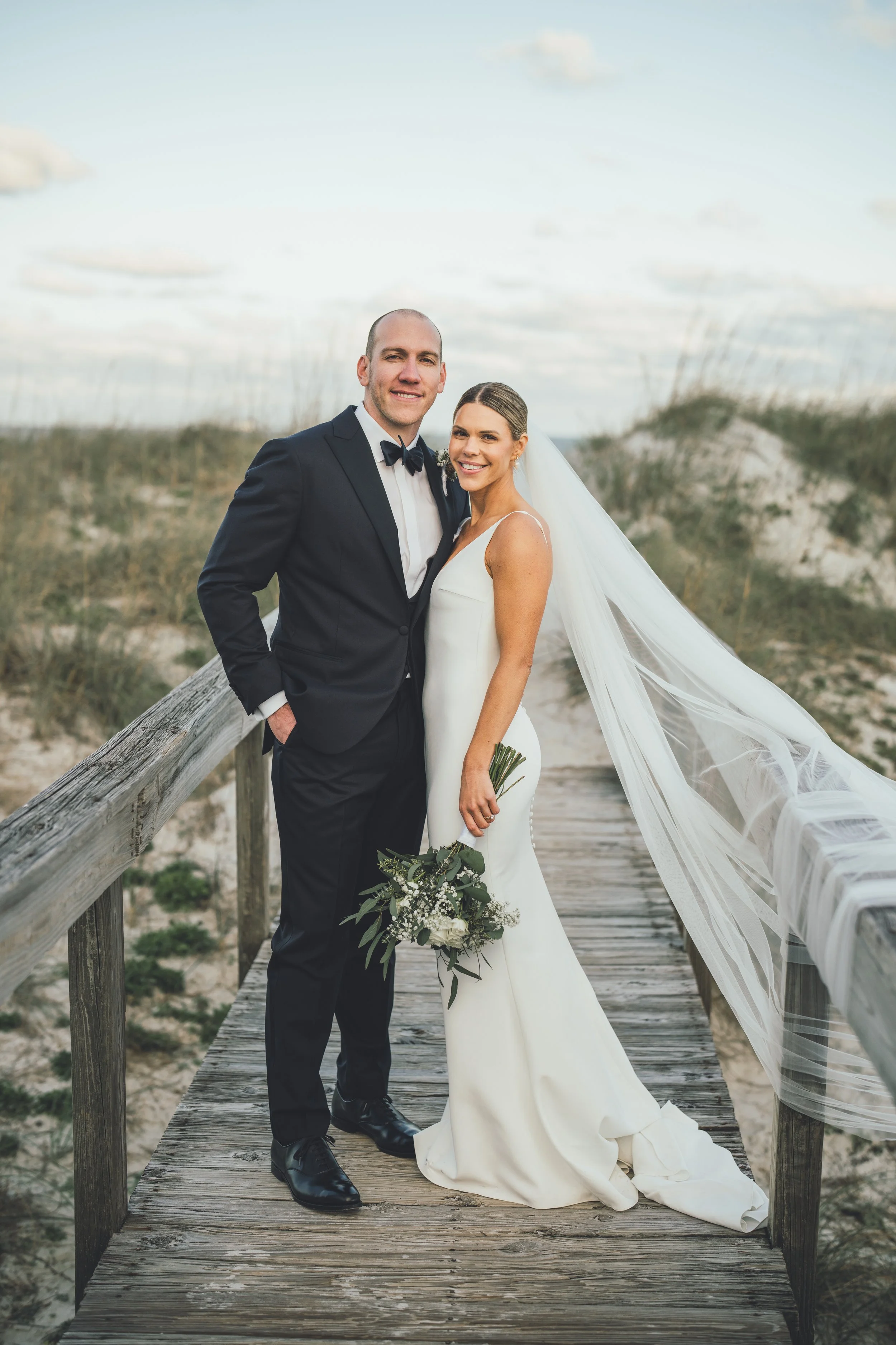 A newly married couple in wedding attire standing on a wooden boardwalk in a beach setting, smiling at the camera.