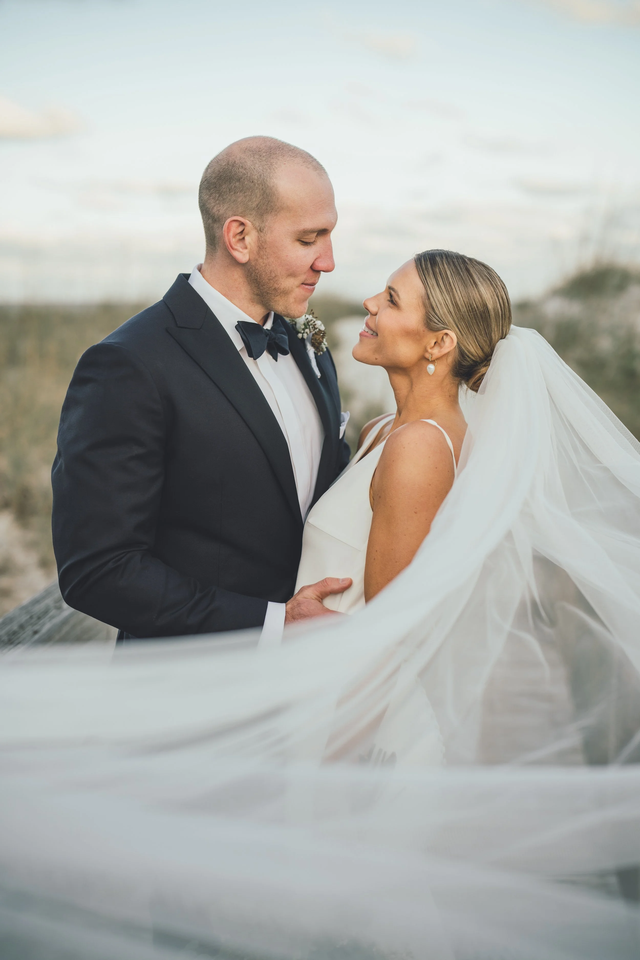 A groom and bride on their wedding day gazing at each other, outdoors with cloudy sky.
