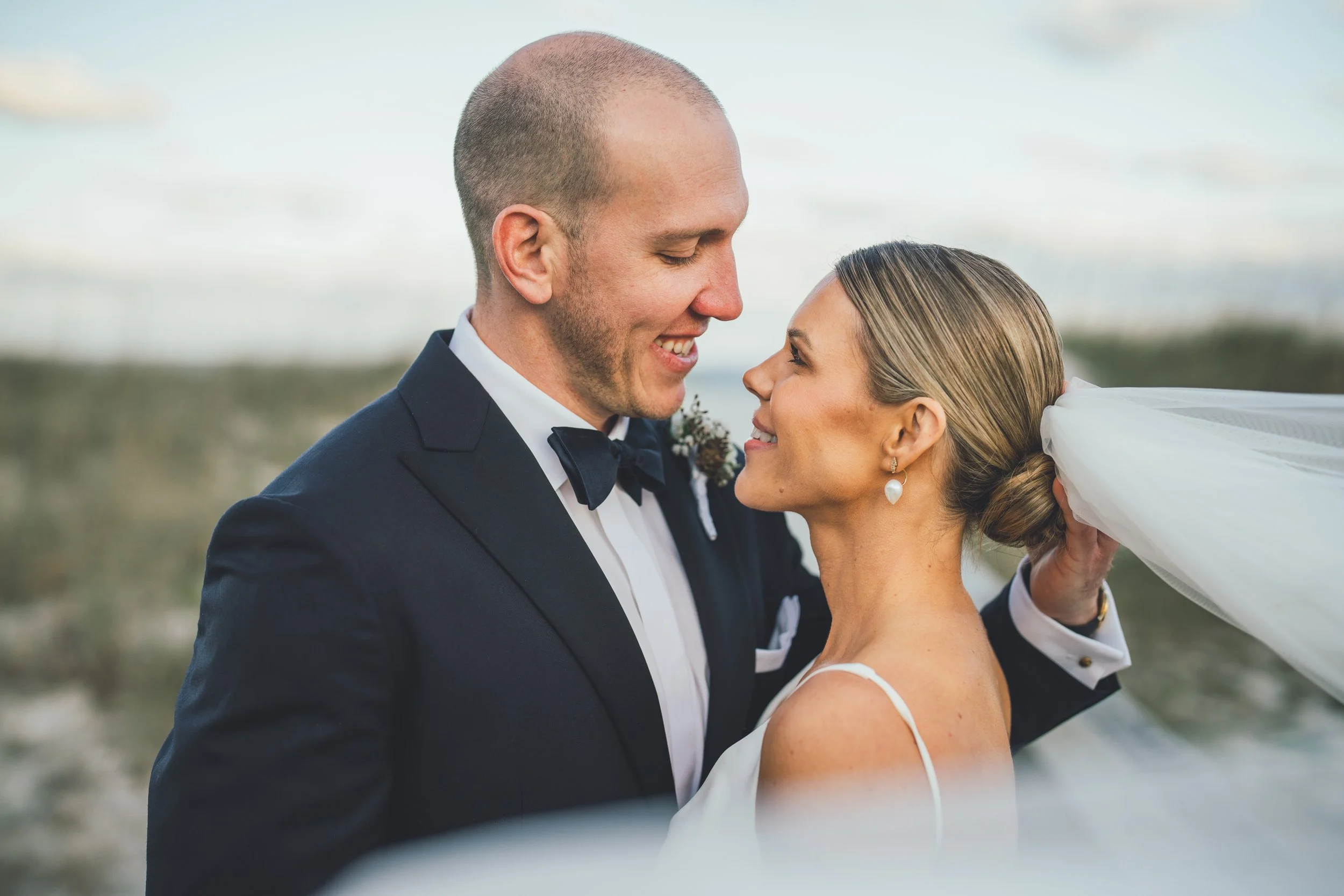 A couple on their wedding day standing close together outdoors, gazing into each other's eyes with smiles, with a blurry natural background and the bride's veil flowing behind her.