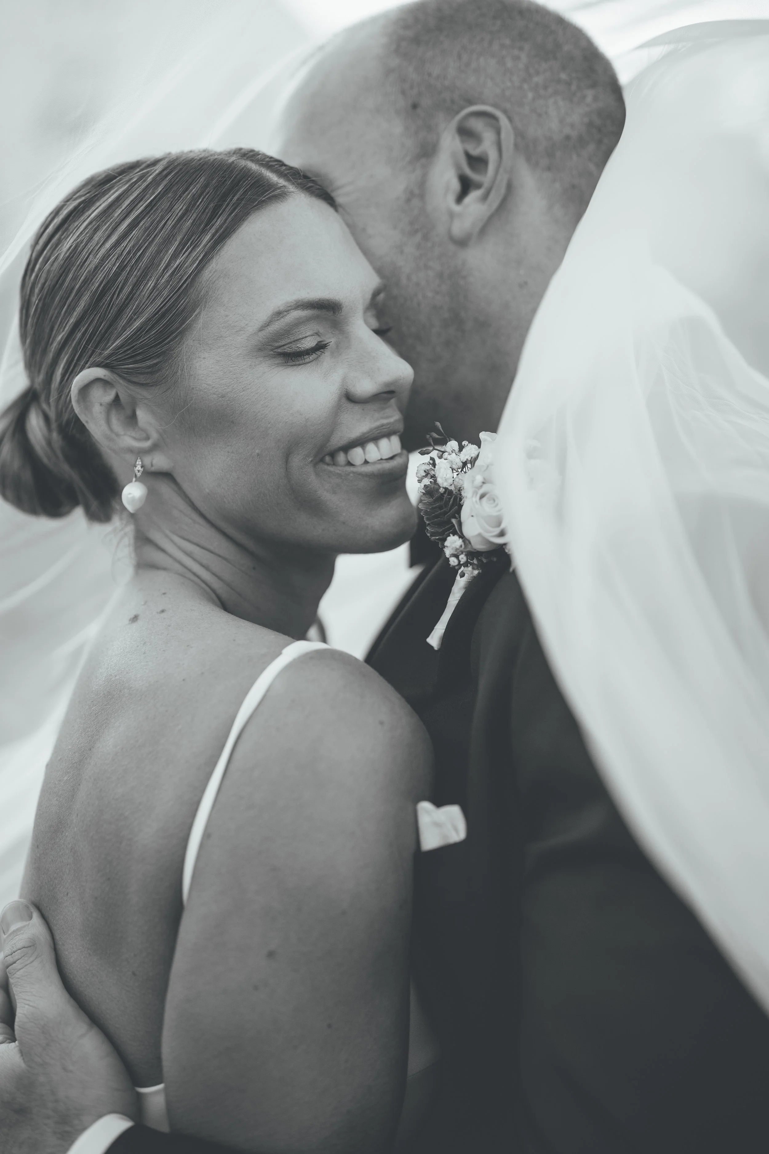 A close-up black and white photograph of a smiling woman in bridal attire, with her eyes closed, leaning into a man who is wearing a tuxedo, both under a wedding veil.