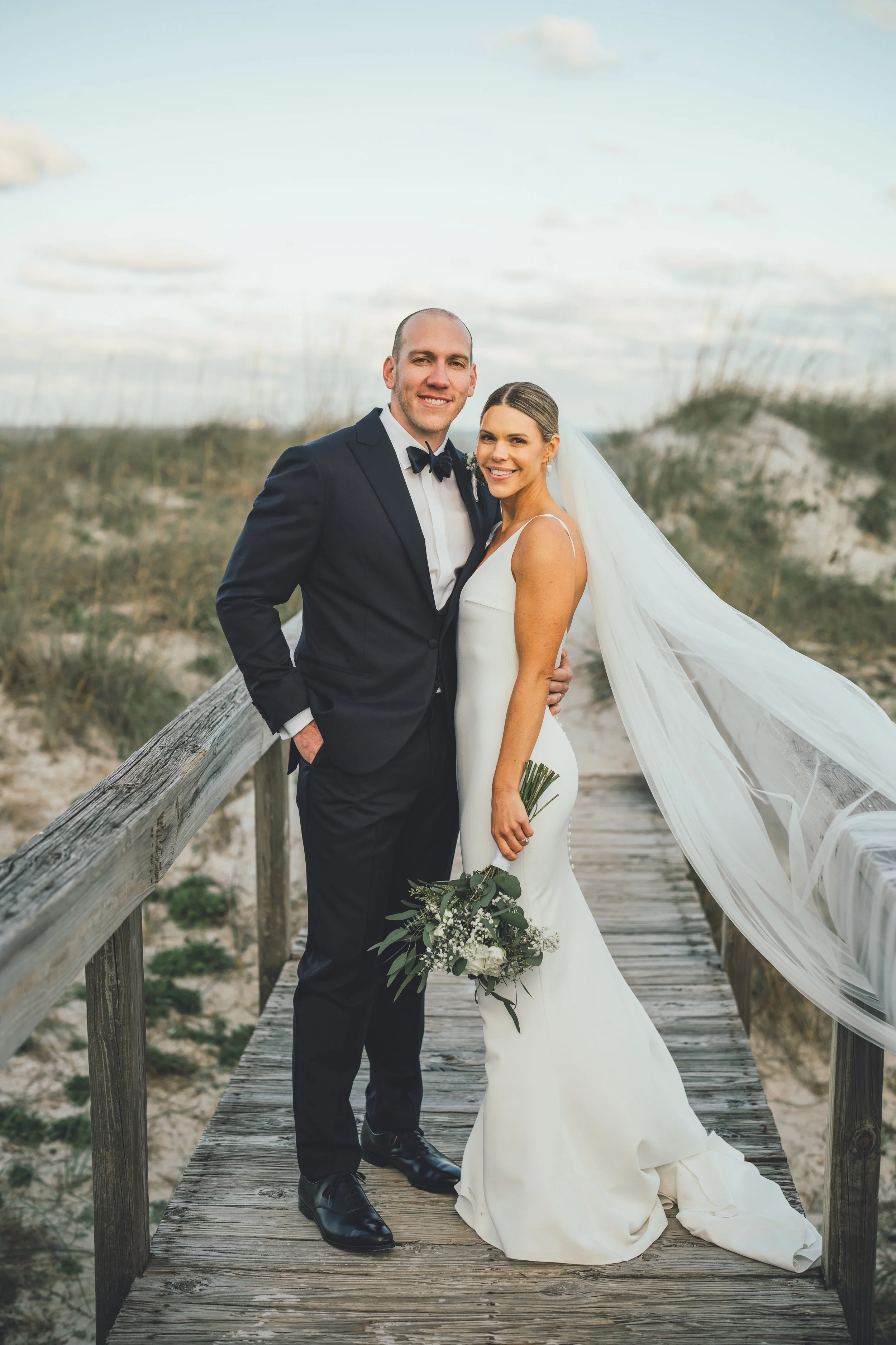 A newlywed couple stands on a wooden boardwalk at the beach, with dunes and tall grass in the background. The groom is in a black tuxedo and the bride in a white wedding gown with a long veil, holding a bouquet of white flowers and greenery.