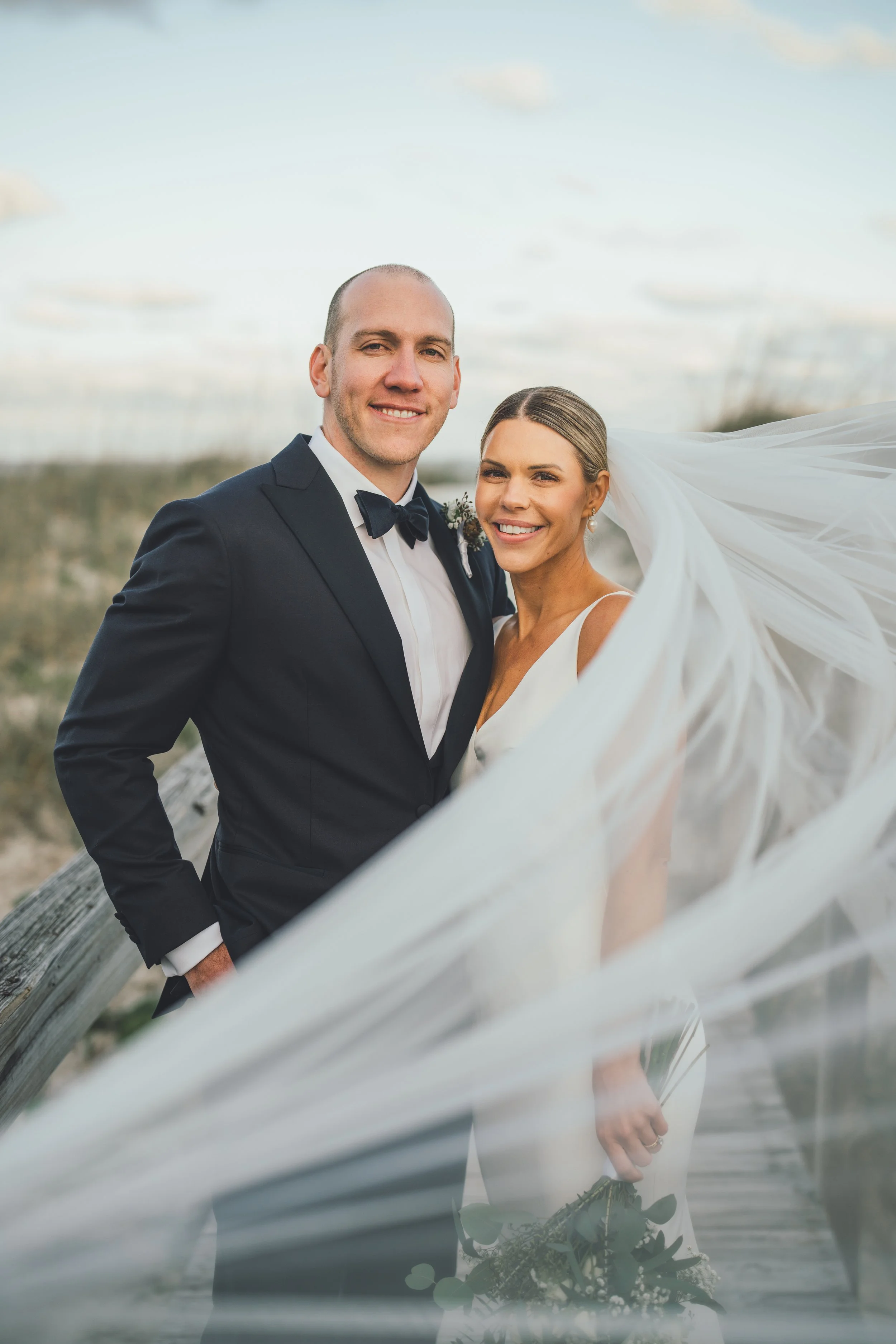 A newlywed couple stands outdoors on a wooden pathway, with the groom in a black tuxedo and bow tie, and the bride in a white wedding dress holding a bouquet, both smiling at the camera against a cloudy sky.