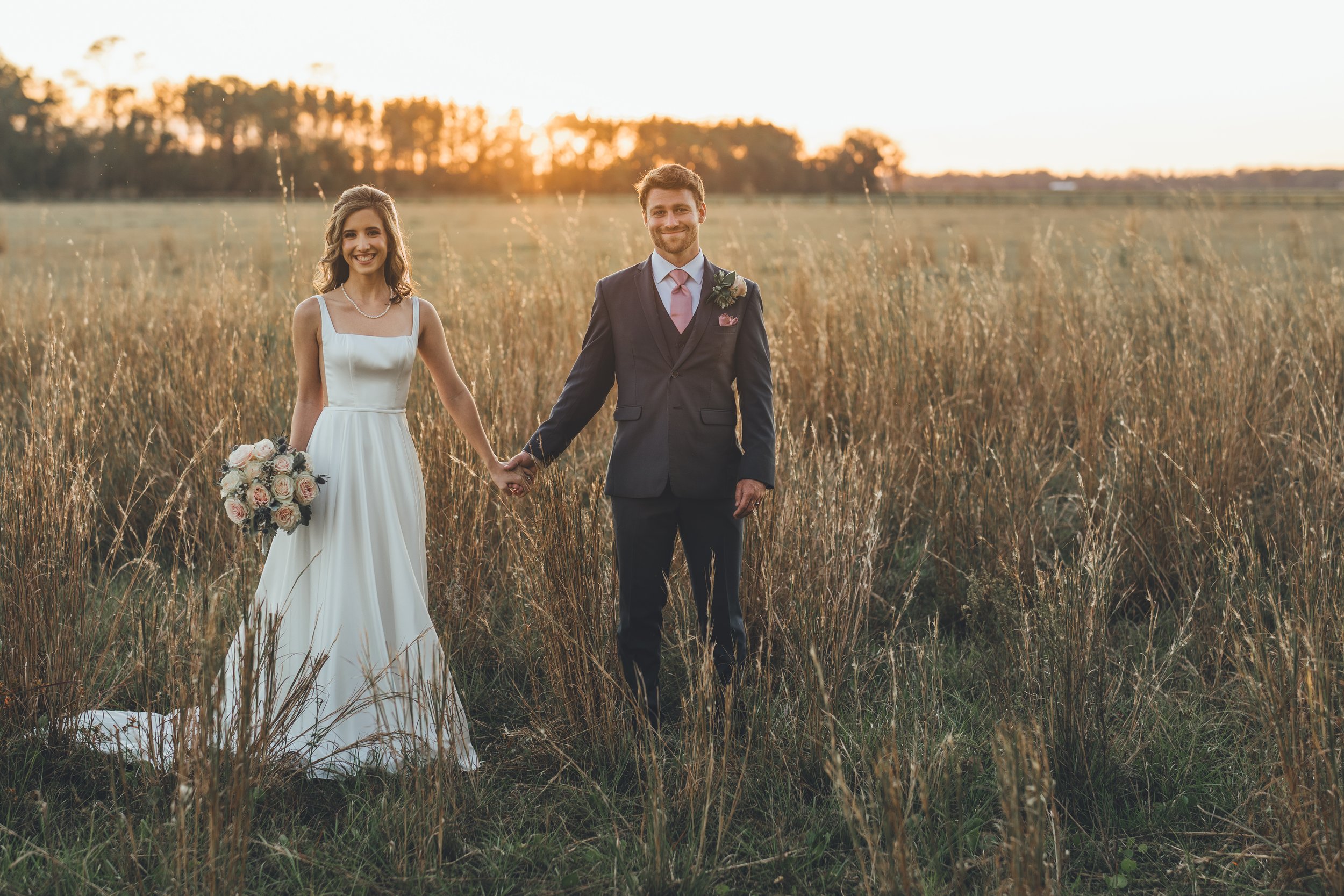 Bride and groom holding hands in a field at sunset, with the bride in a white wedding gown holding a bouquet of flowers, and the groom in a dark suit with a pink tie and boutonniere.