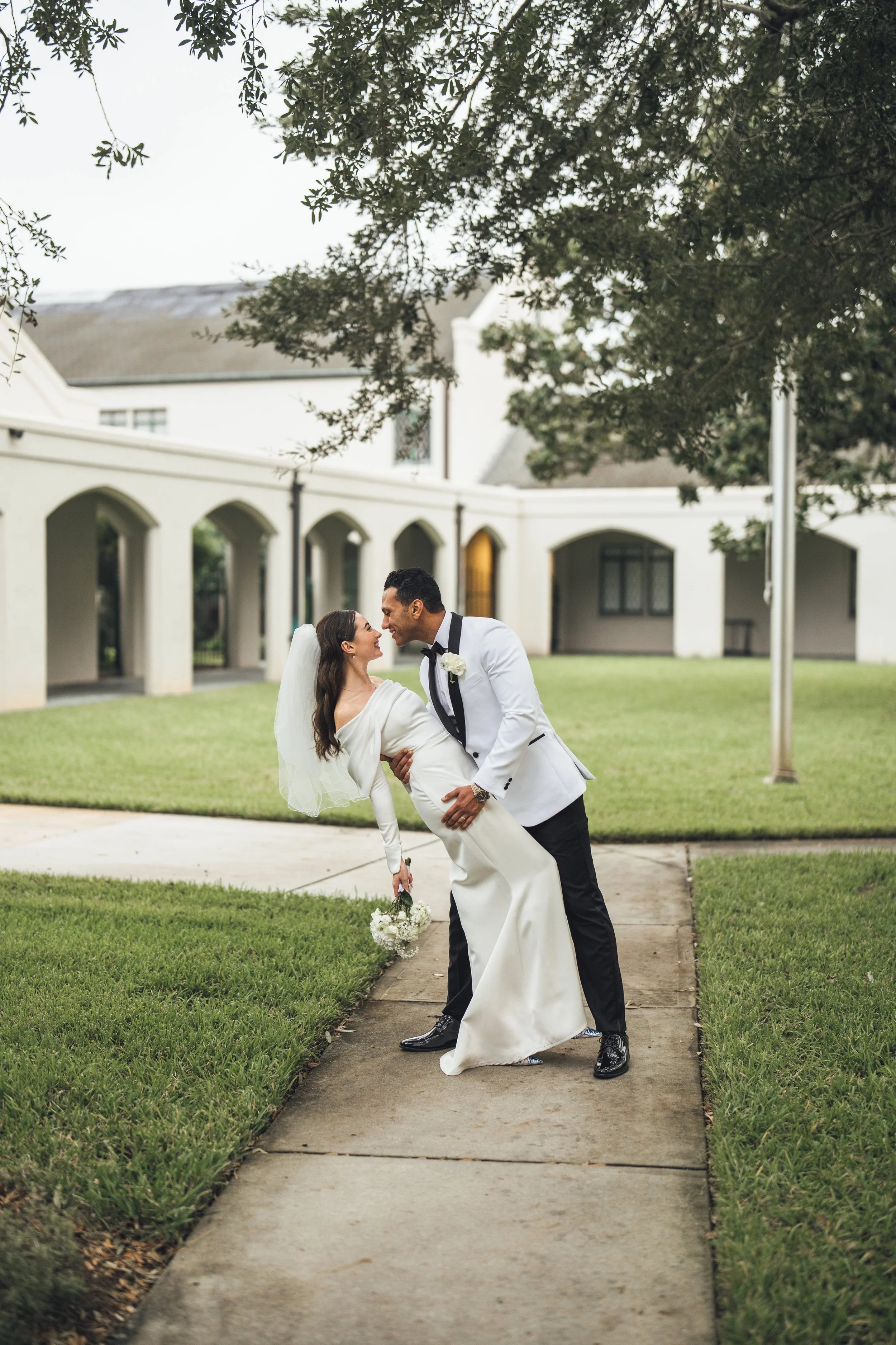 A bride and groom dancing outside on a concrete pathway in front of a white building with arched doorways and green lawn, celebrating their wedding.