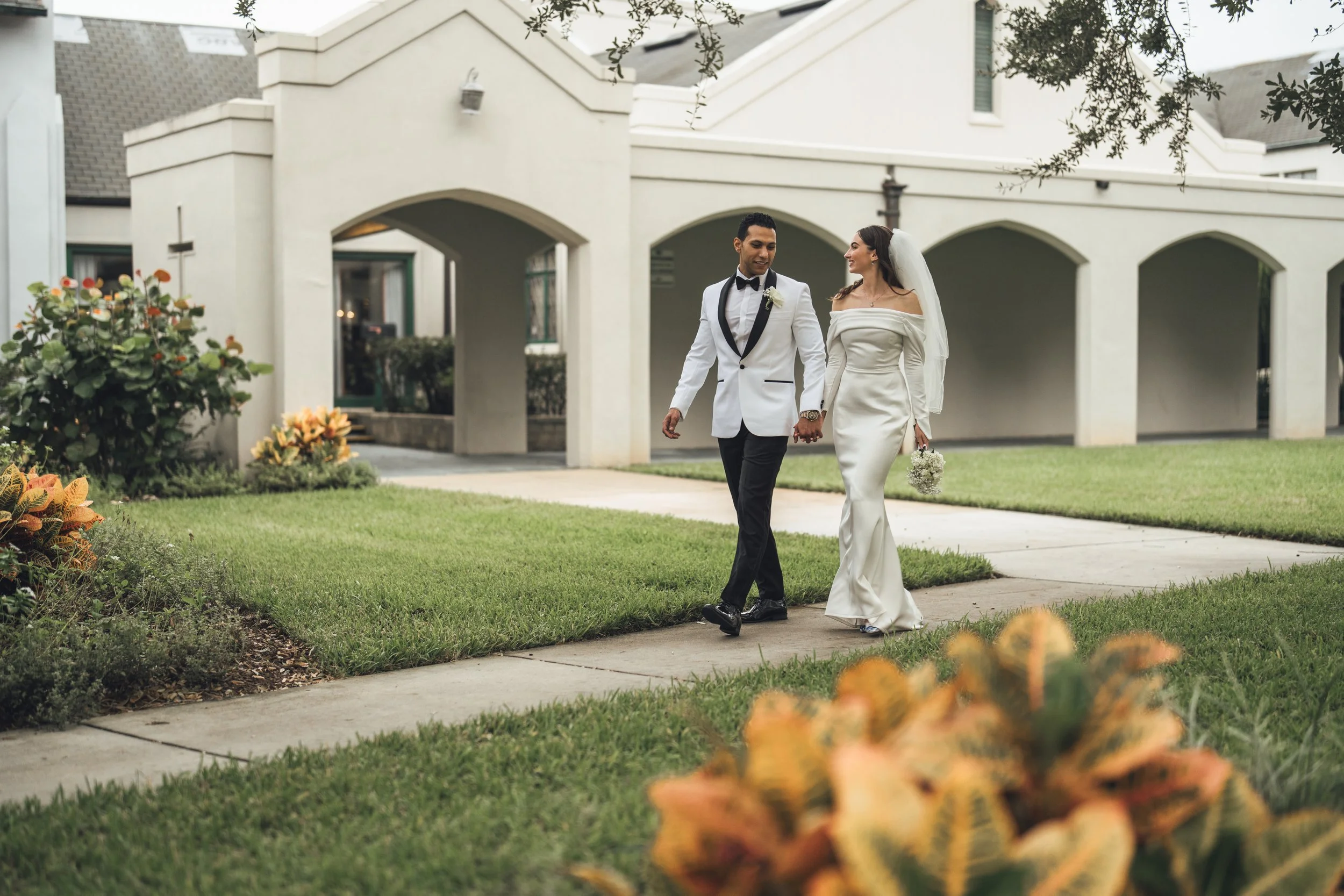 A newlywed couple walking hand in hand on a sidewalk in front of a white building with arches, greenery, and flowers, outdoors during daytime.