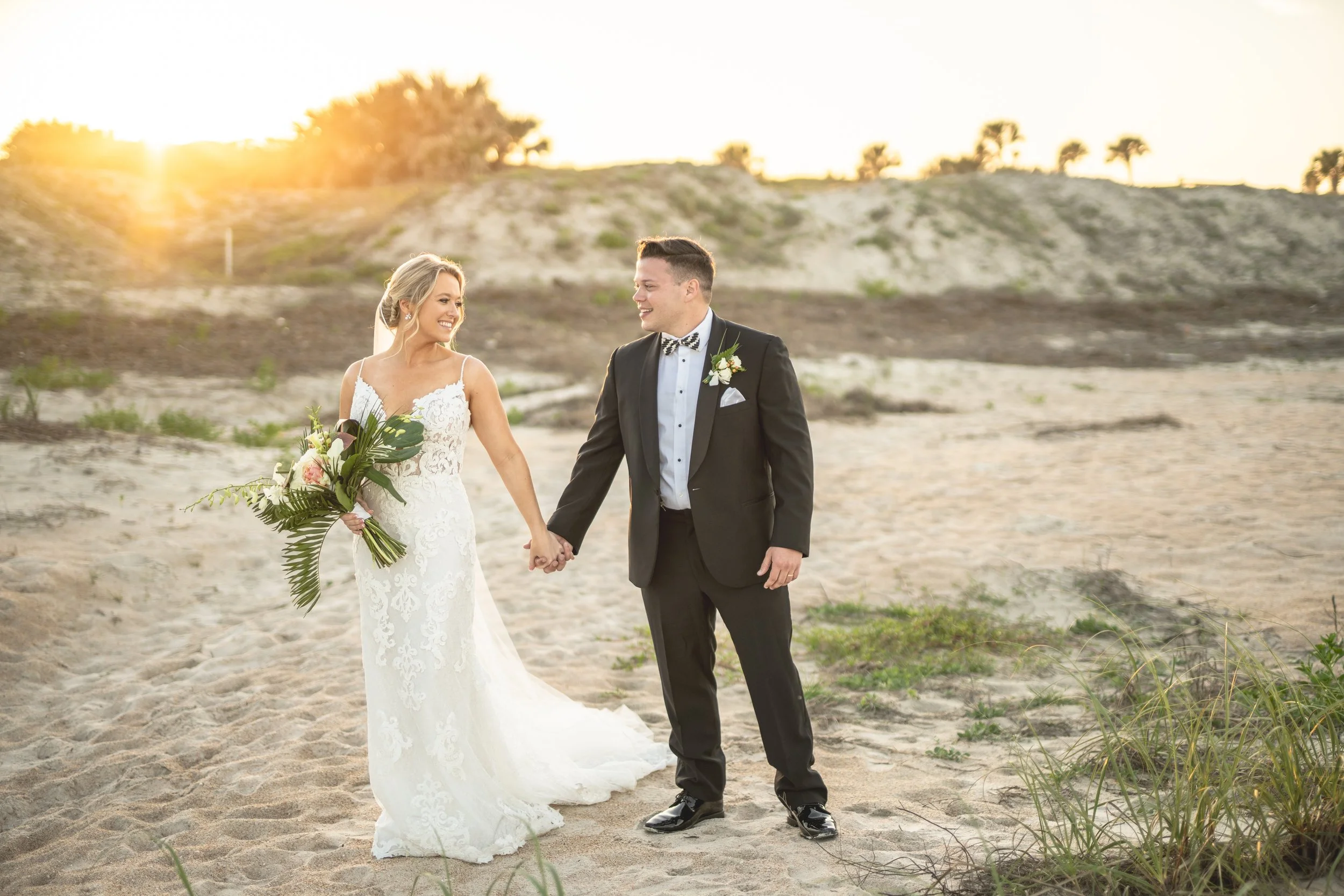 Bride and groom in wedding attire holding hands on a sandy beach at sunset, smiling at each other.