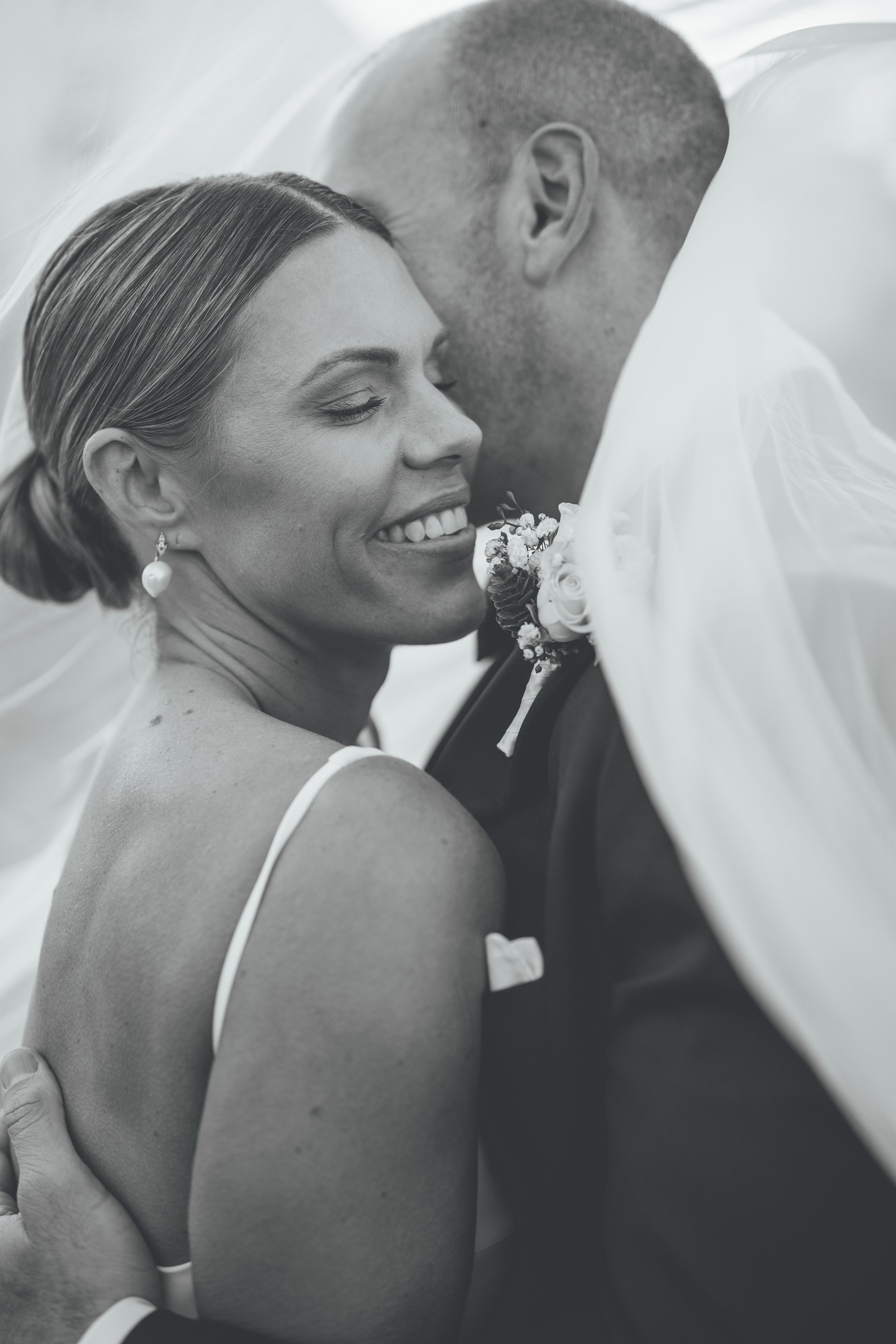Black and white photo of a bride and groom sharing a tender moment, with the bride smiling and the groom leaning in close, wearing wedding attire, wedding bouquet on his lapel, veil partially covering the bride's face.