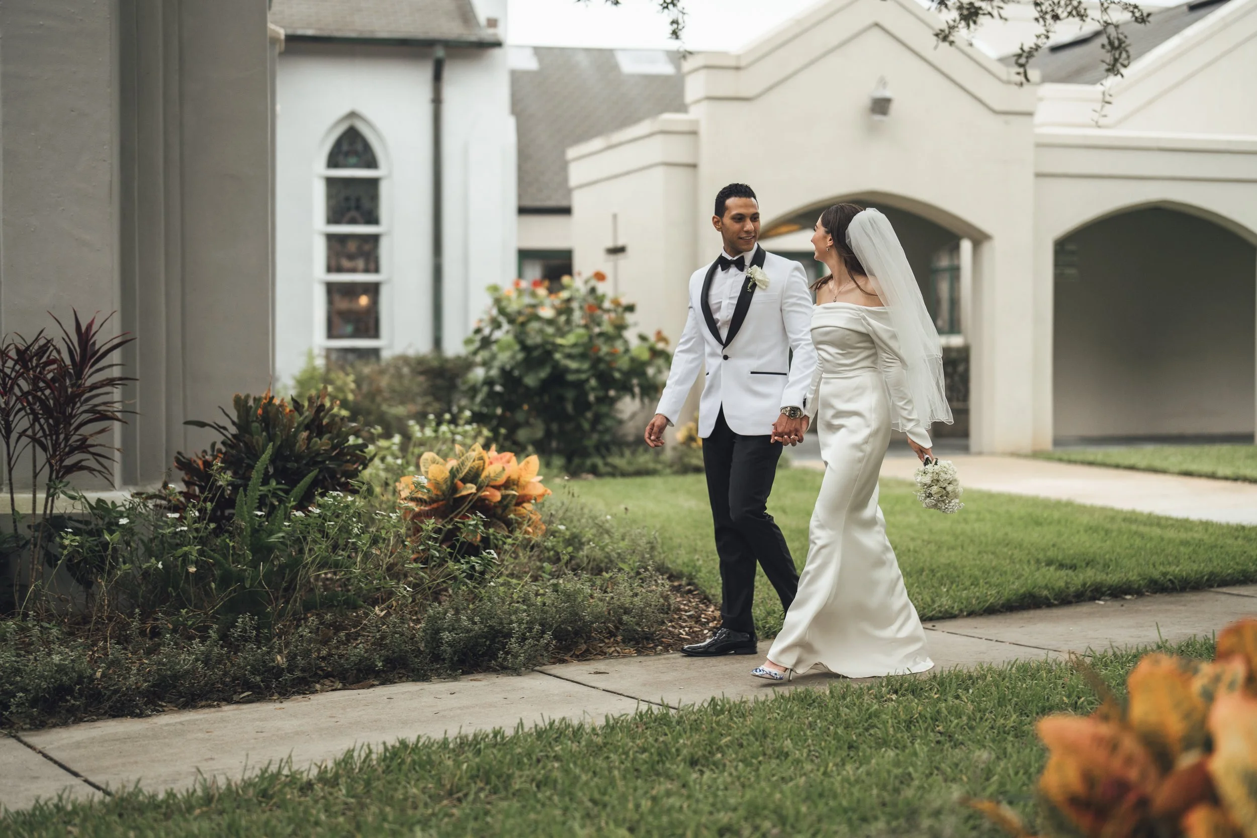 A couple dressed in wedding attire, holding hands and walking outside near a church, with flowers and greenery around them.