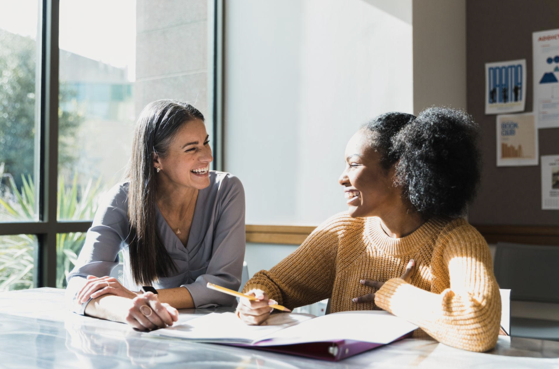 Two women smiling and discussing notes at a sunlit office table.