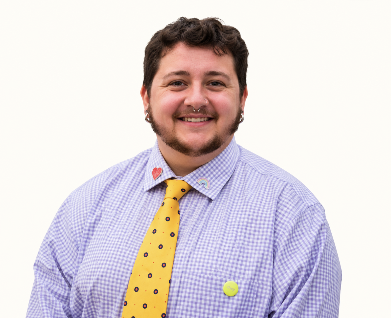 Smiling person wearing a lavender gingham shirt, yellow patterned tie, and colorful rainbow and heart pins
