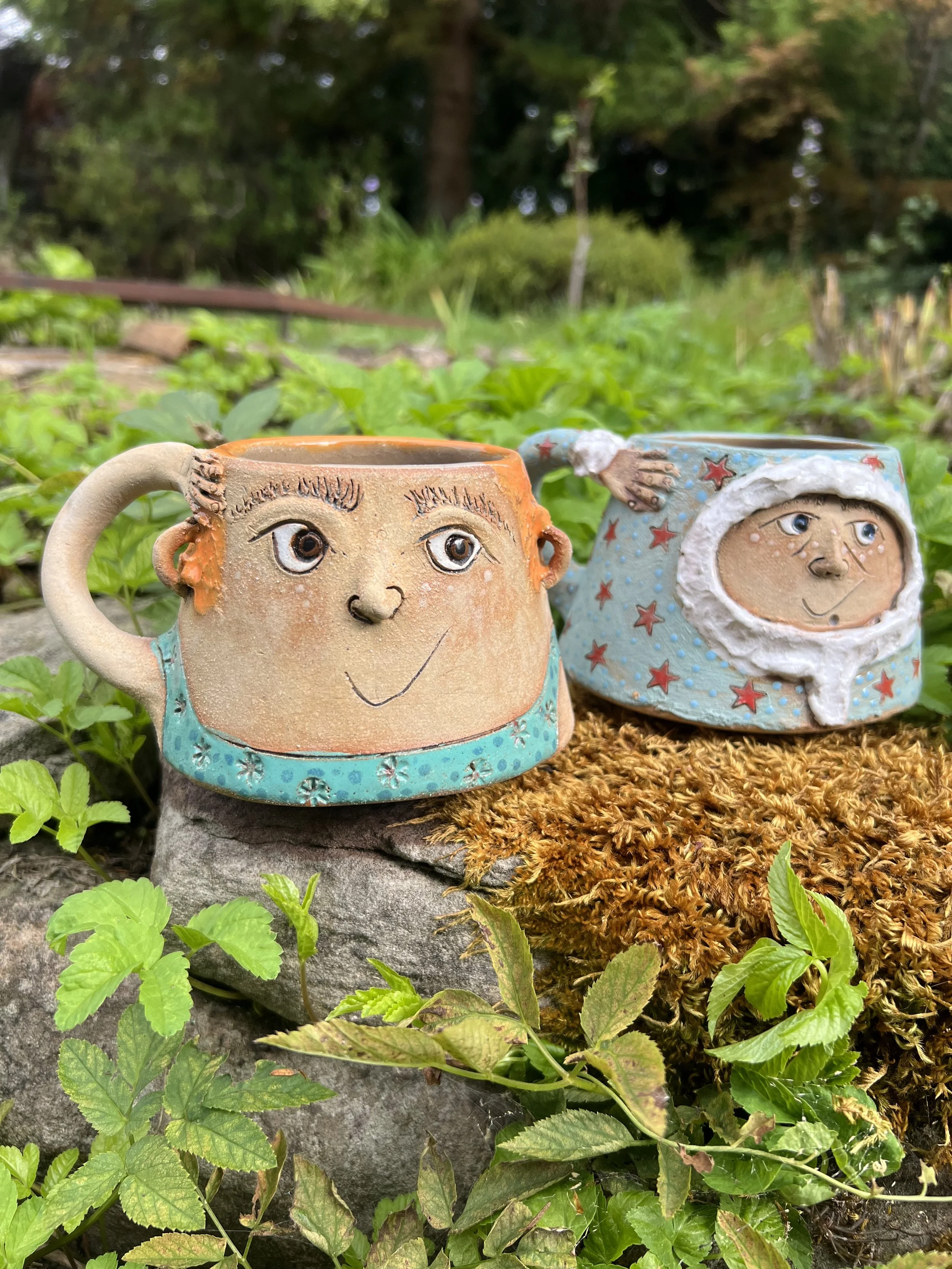 Two whimsical ceramic mugs with faces, one resembling a person with short hair and the other dressed in a puffer coat, placed on a rock amidst greenery and moss in an outdoor setting.