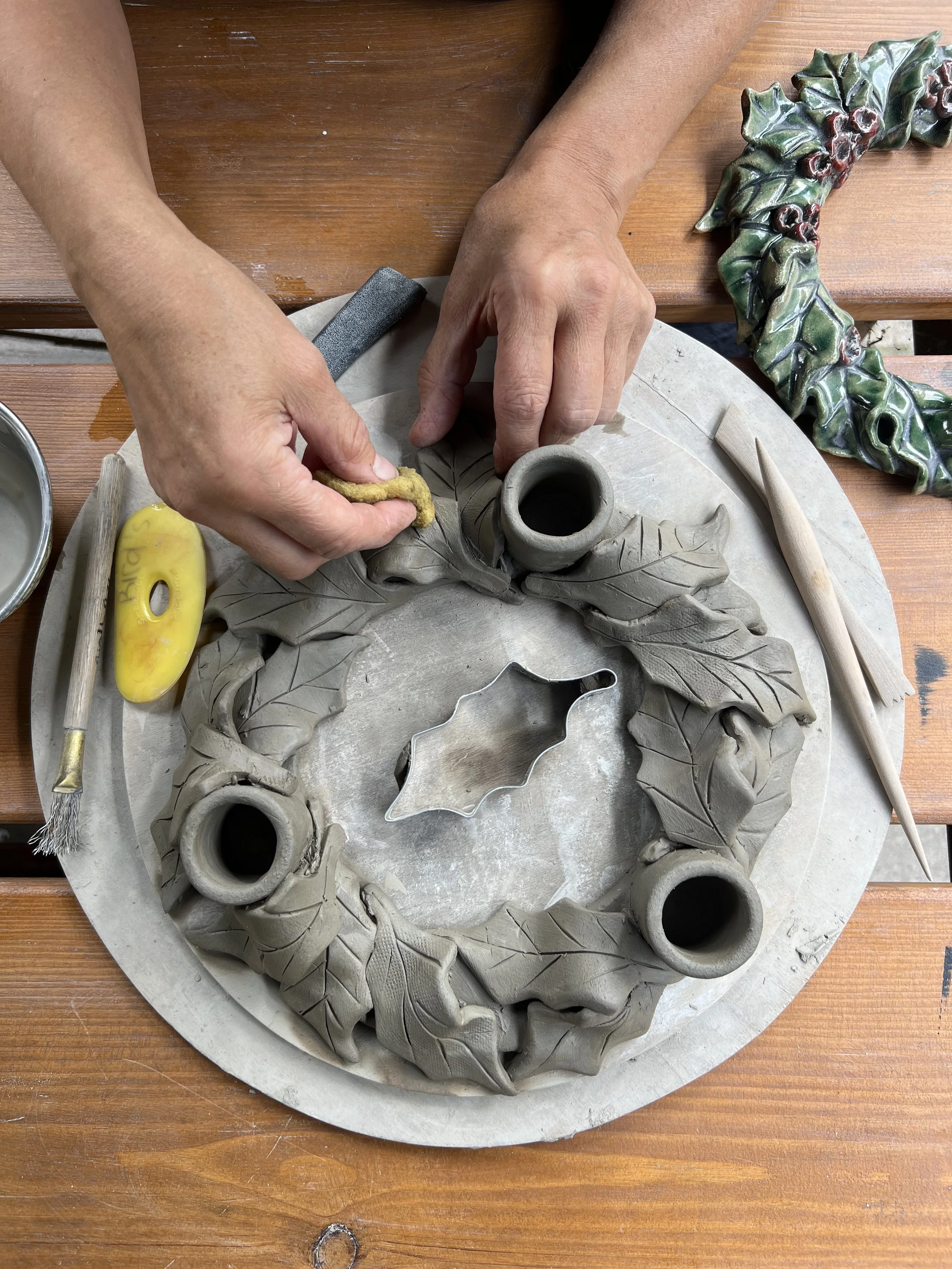 People are working on a clay sculpture of a wreath with leaves and holly berries on a wooden table.